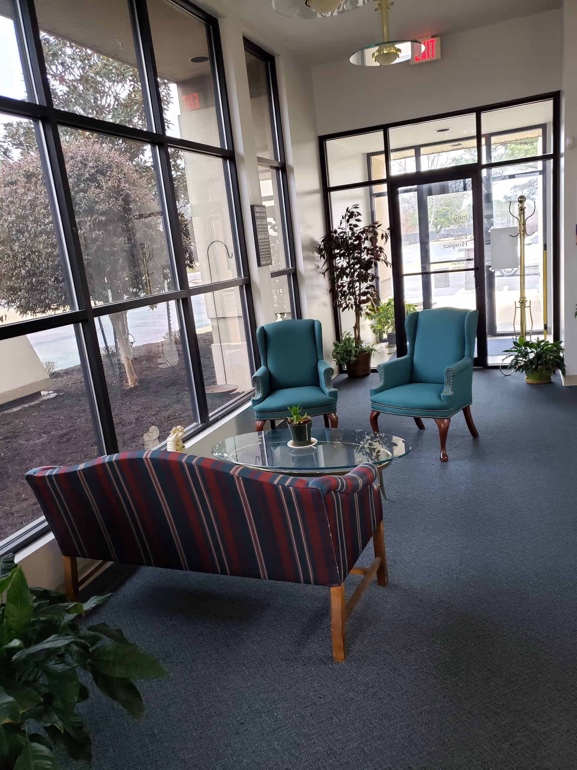 Bright lounge area in a facility with teal chairs, striped sofa, glass coffee table, plants.