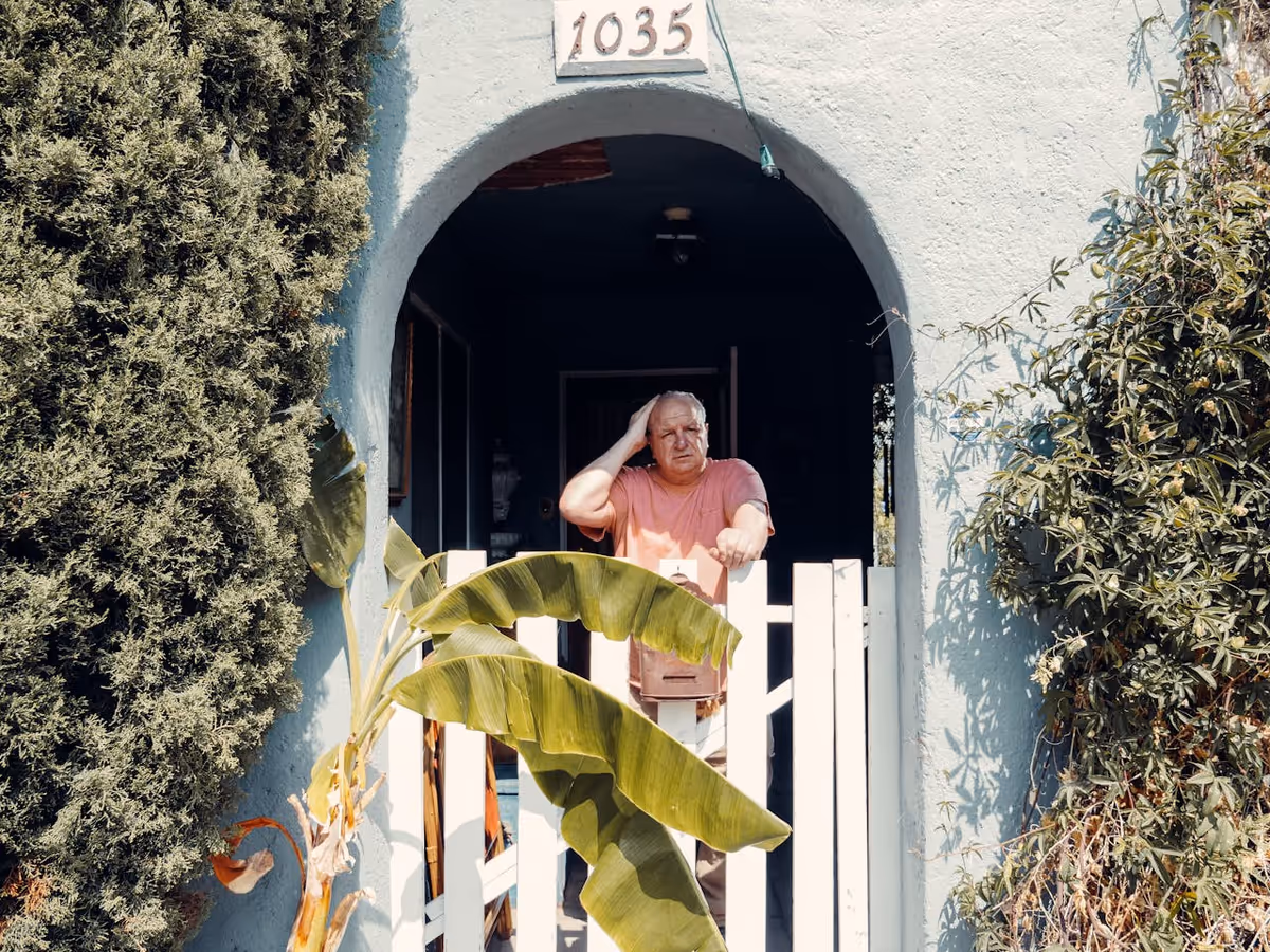 Elderly man standing at a blue arched doorway with a white gate and greenery.
