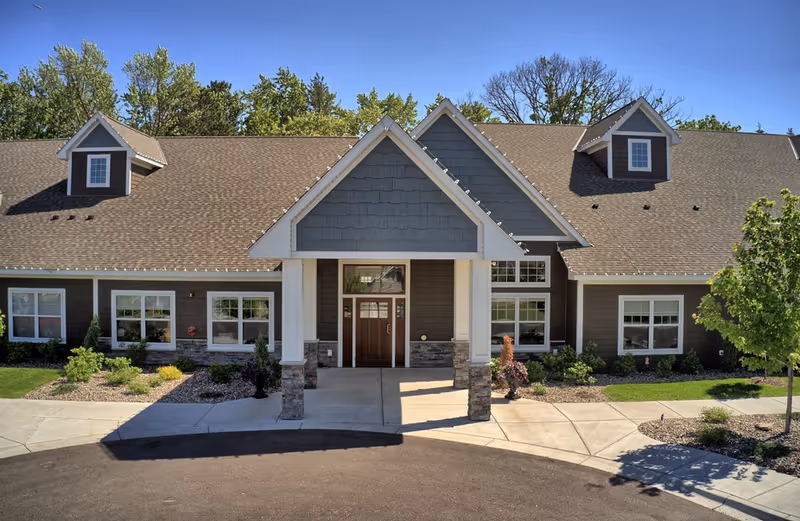 Front entrance of a single-story senior living facility with a gabled roof, columns, landscaping and a curved driveway.