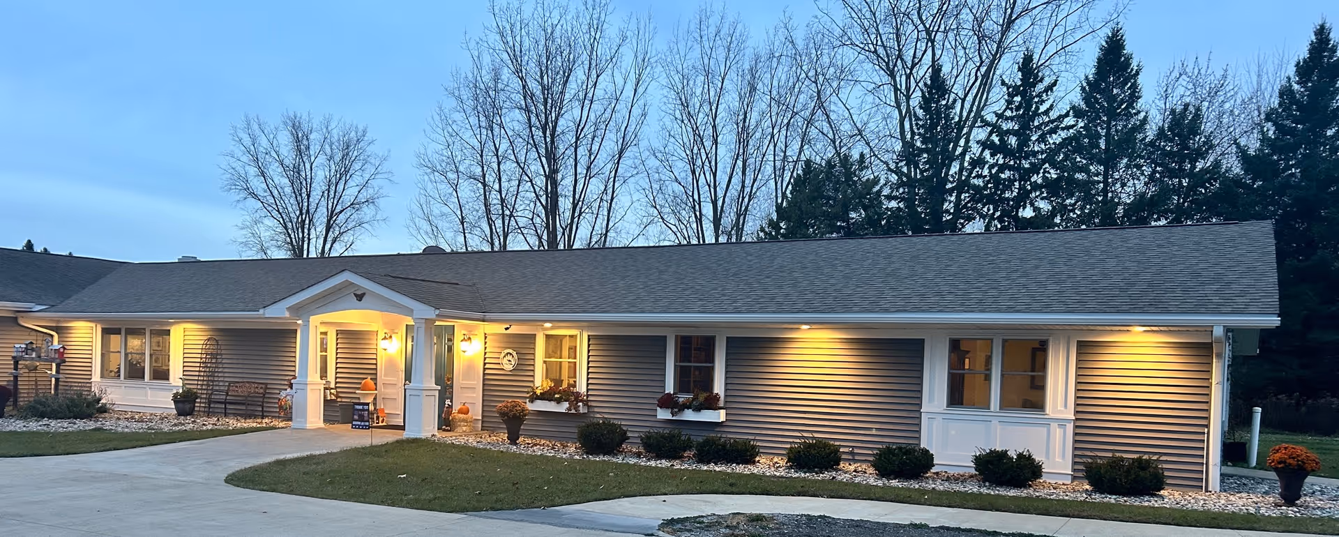 Single-story senior living building with a lit front entrance, shrubs, and a curved driveway at dusk.