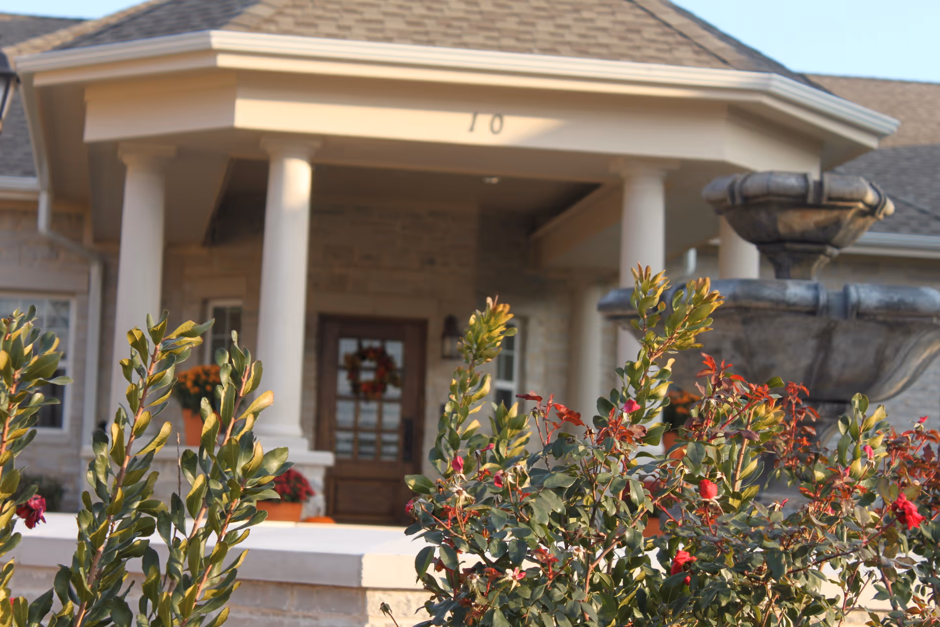 Entrance of a senior living facility with a covered porch supported by white columns, a wooden door decorated with a wreath, and a large stone fountain to the right. Green and red foliage is visible in the foreground.