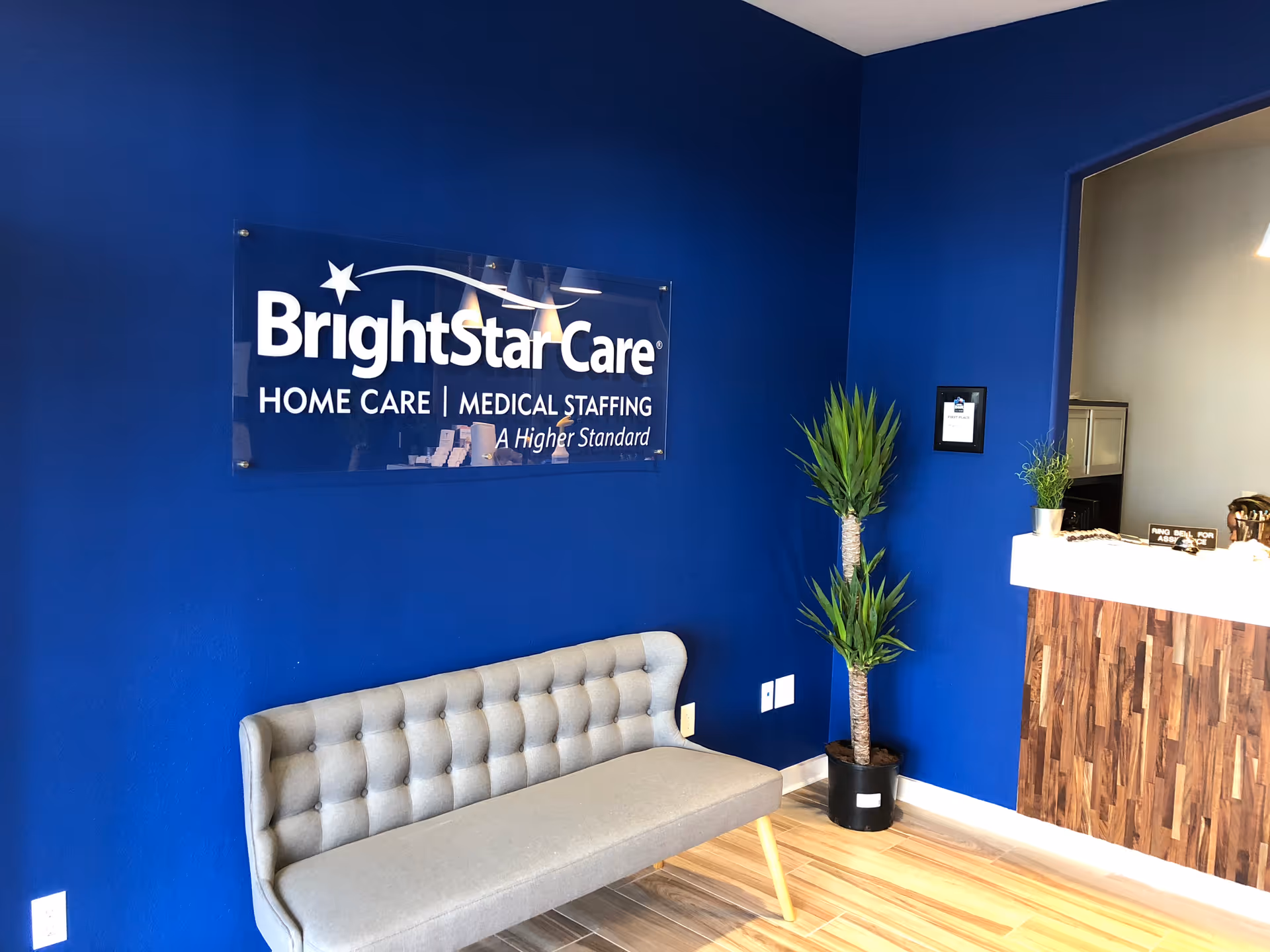 Blue-walled reception area with BrightStar Care sign, gray tufted couch, and tall plants.