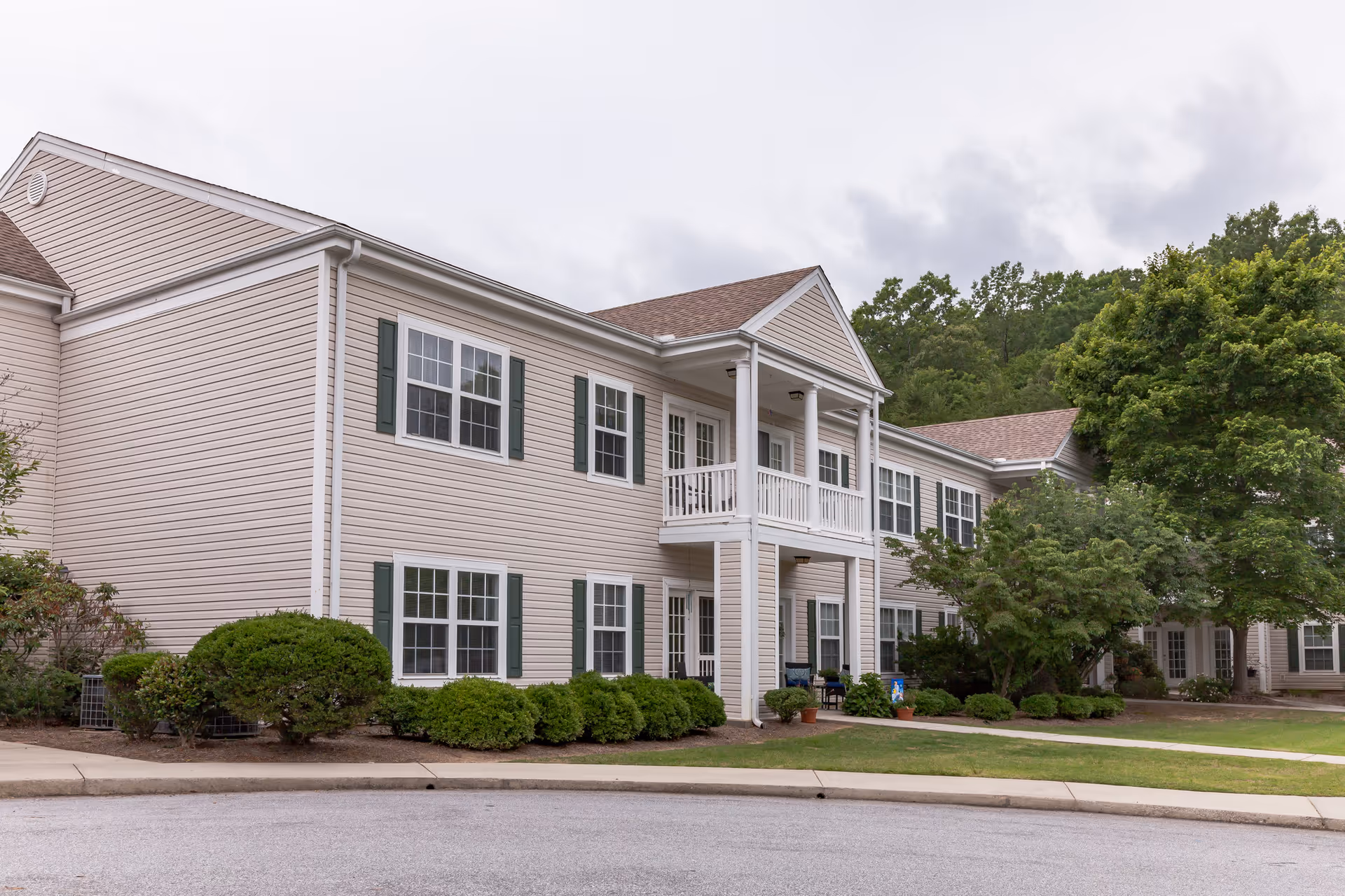Two-story beige siding building with a covered entrance and balcony, surrounded by shrubs and a lawn.