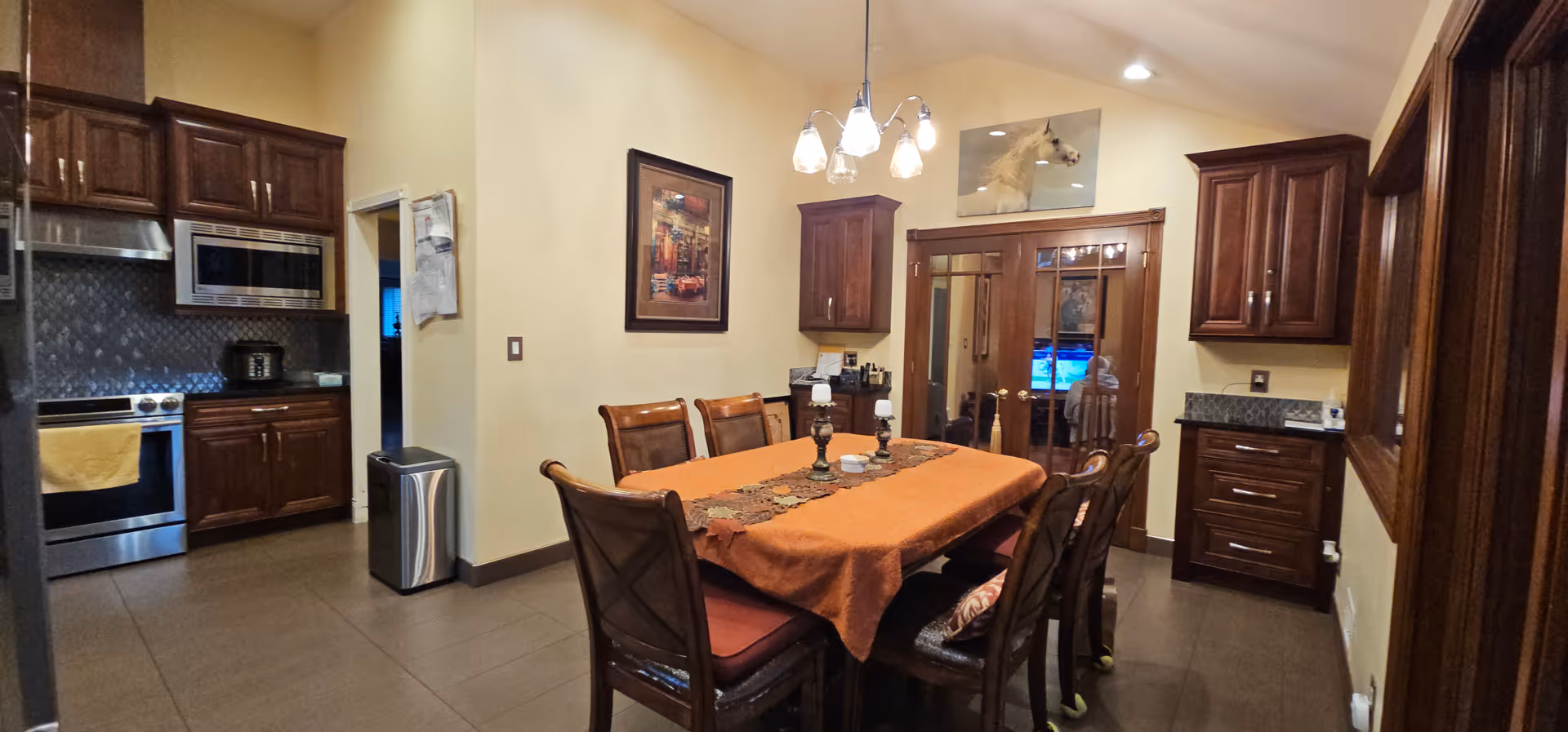 A formal dining room with a long table, chairs, chandelier, and a view into the kitchen.