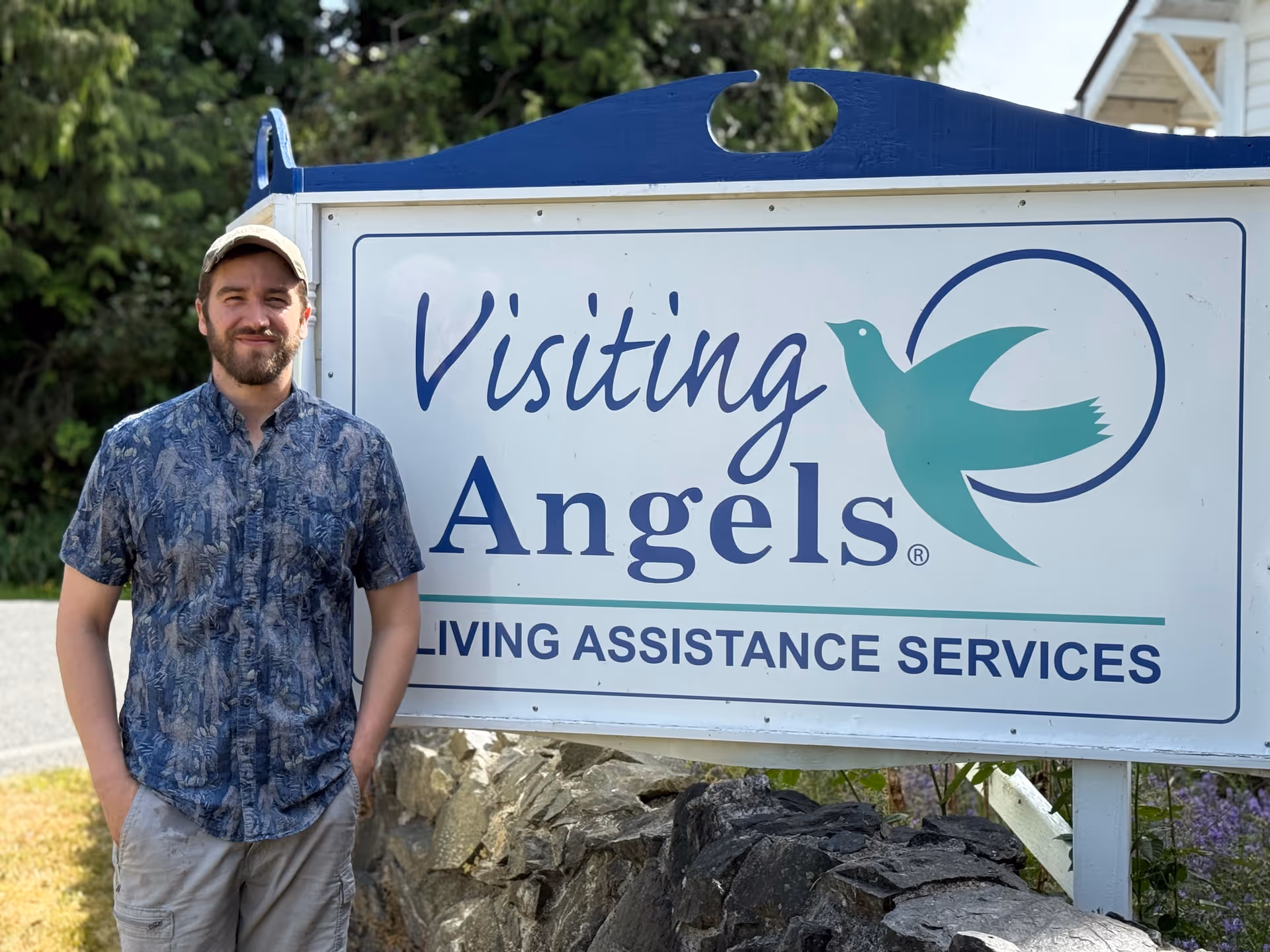 Man stands beside Visiting Angels sign outdoors advertising senior home care services.