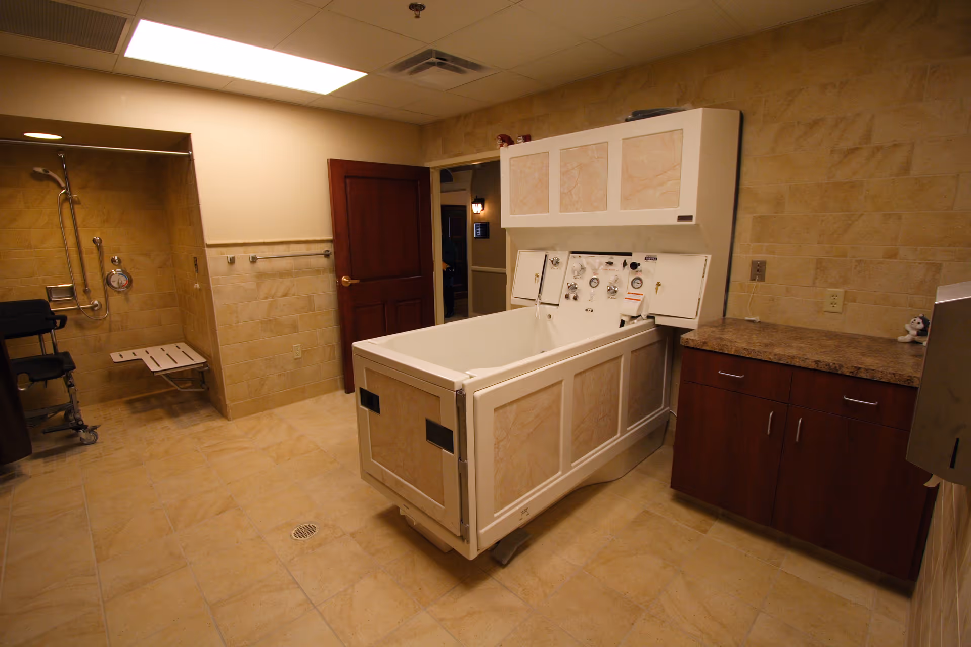 A spacious bathroom with beige tiled walls and floor, featuring a large white therapeutic bath tub with control panels, a shower area with a foldable seat and grab bars, a wooden door, and a countertop with cabinets underneath.