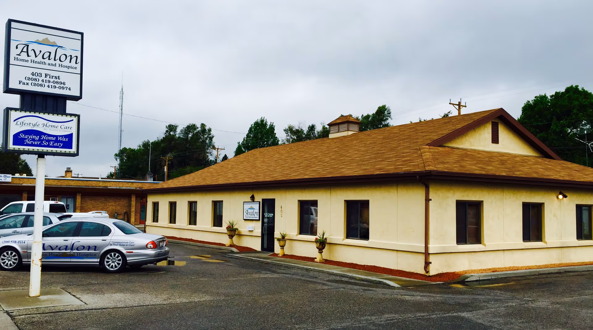 A single-story beige building with a brown roof, signs, and parked cars in a lot.