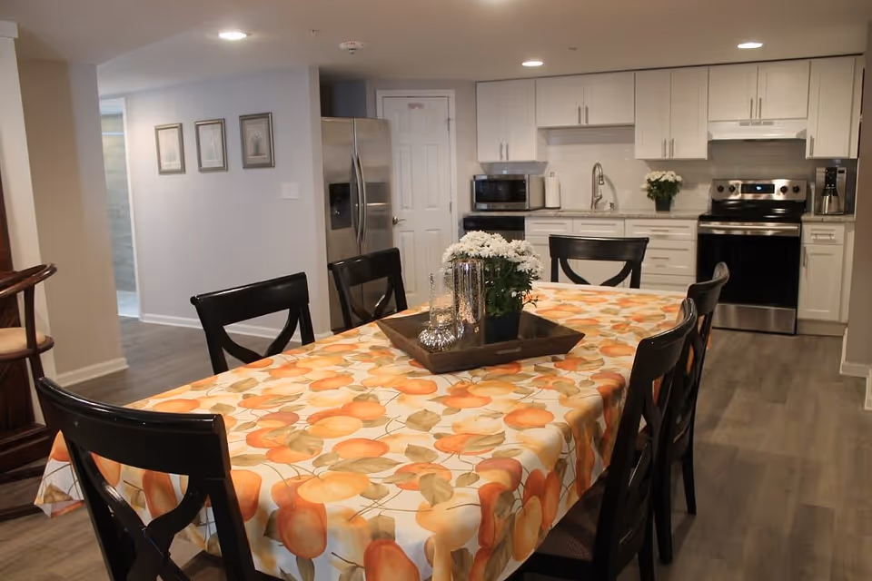 A dining area with a rectangular table covered with a tablecloth featuring an orange fruit pattern. The table is surrounded by six black chairs. In the background, there is a modern kitchen with white cabinets, a stainless steel refrigerator, stove, microwave, and a coffee maker. A small potted plant and decorative items are placed on the table.