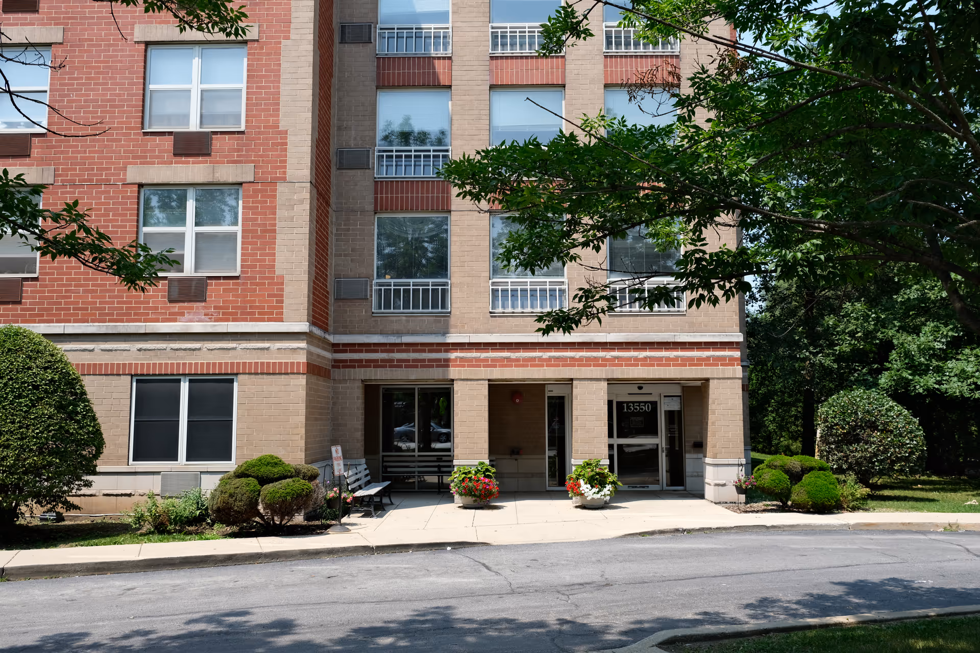 Front entrance of a brick senior living building with potted flowers, benches, and the address 13550 visible on the door.