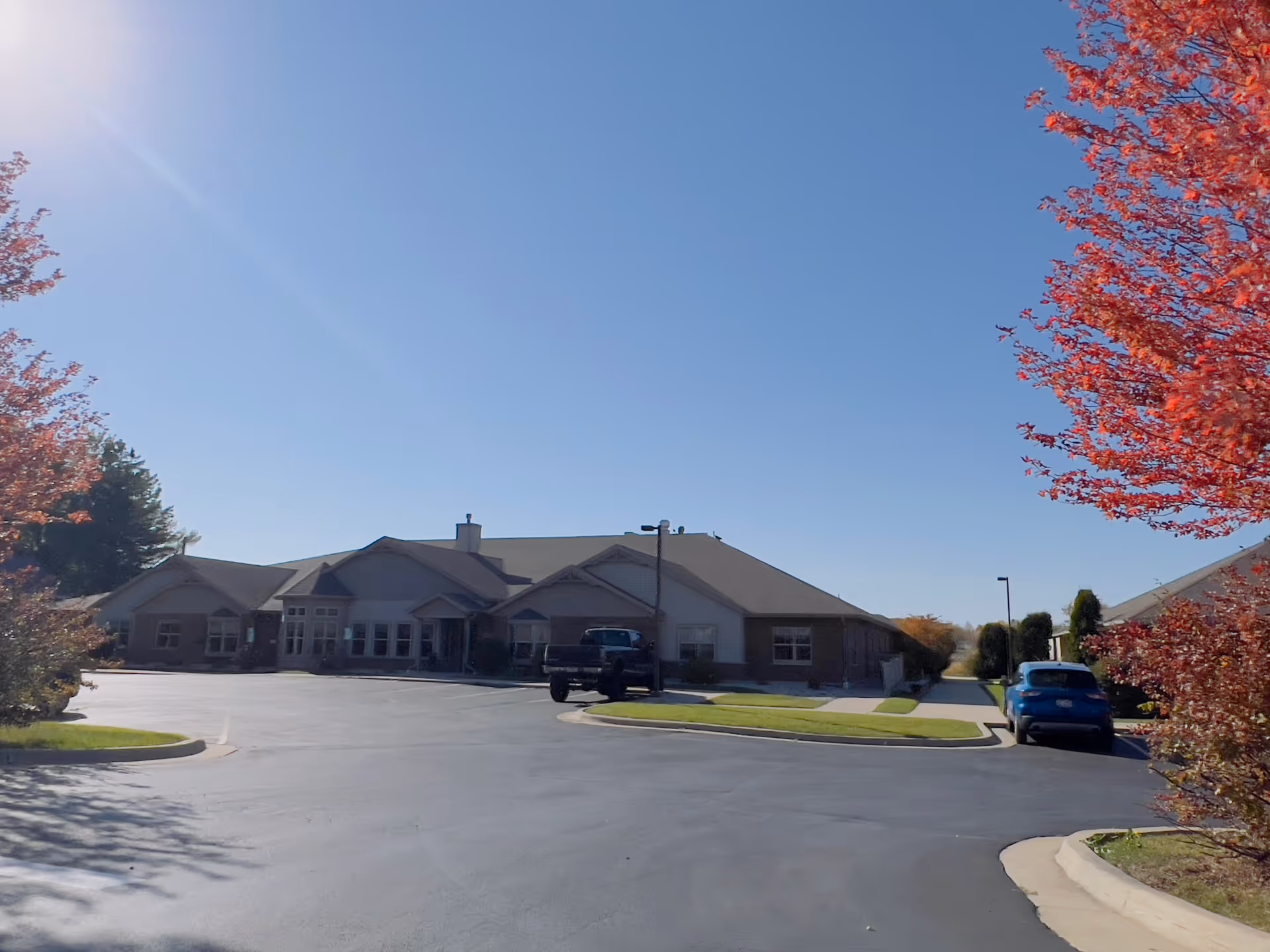 Single-story senior living facility building with a parking lot, cars, and red-leaved trees under a clear blue sky.