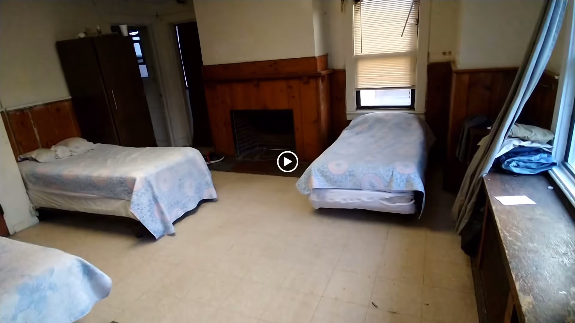 A simple bedroom with three single beds covered with light blue patterned bedspreads. The room has wood paneling on the walls, a fireplace in the center, a wardrobe in the corner, and a window with blinds partially drawn. There is a wooden ledge under the window with some folded clothes and a piece of paper on it.