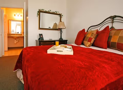 Cozy bedroom with a red quilted bedspread, decorative pillows, and a dresser against the wall.