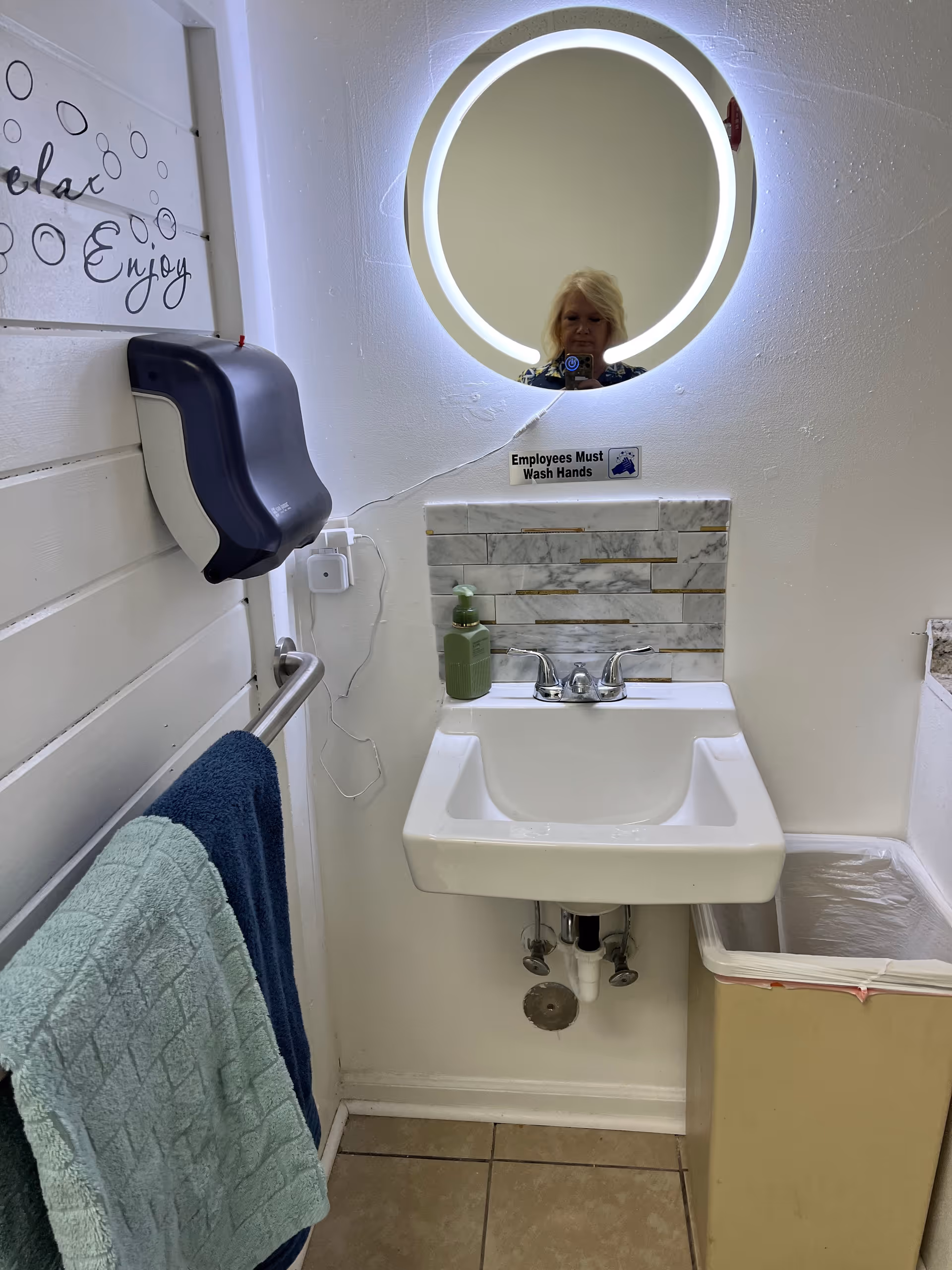 Small bathroom with a round backlit mirror, sink, towels, and an 'Employees Must Wash Hands' sign.