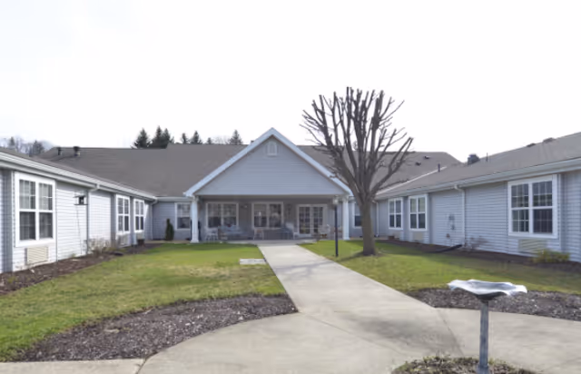 Exterior view of a single-story senior living facility building with a central covered porch area, surrounded by a lawn and a concrete walkway leading to the entrance. There is a leafless tree and a birdbath in the courtyard area.