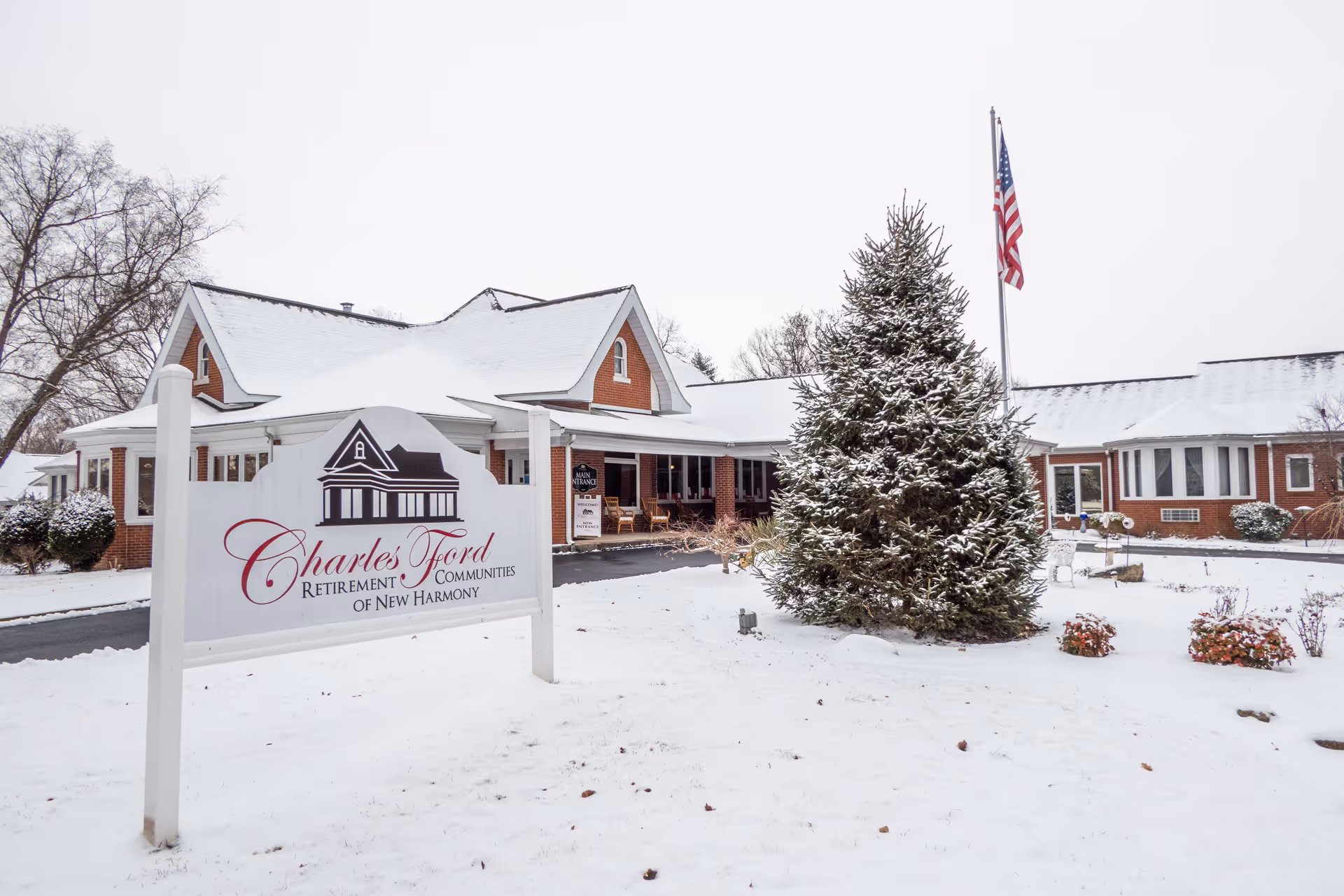 Snow-covered exterior of Charles Ford Retirement Communities of New Harmony building with a large sign in the foreground, an American flag on a flagpole, and a snow-dusted evergreen tree.