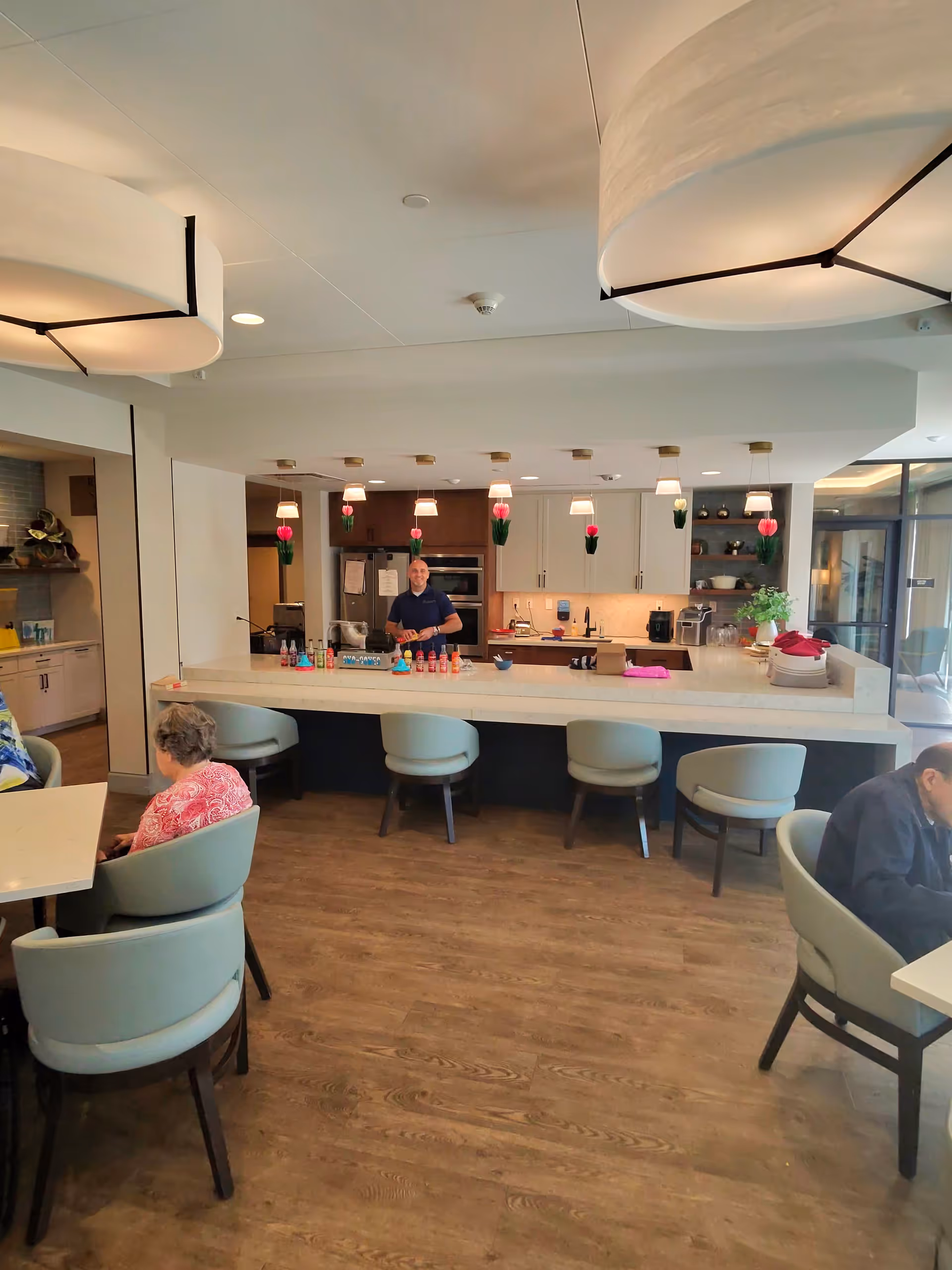 Staff member serves residents at a bar-style counter in a bright dining area.