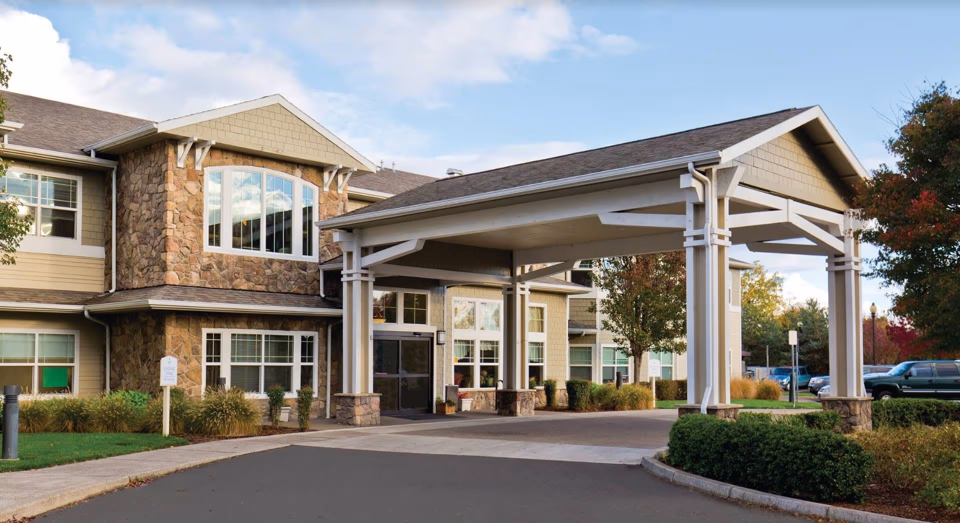 Exterior view of Meadowlark Senior Living facility showing a two-story building with stone and beige siding, large windows, and a covered entrance driveway. There are trees and shrubs around the building and a parking area visible in the background.