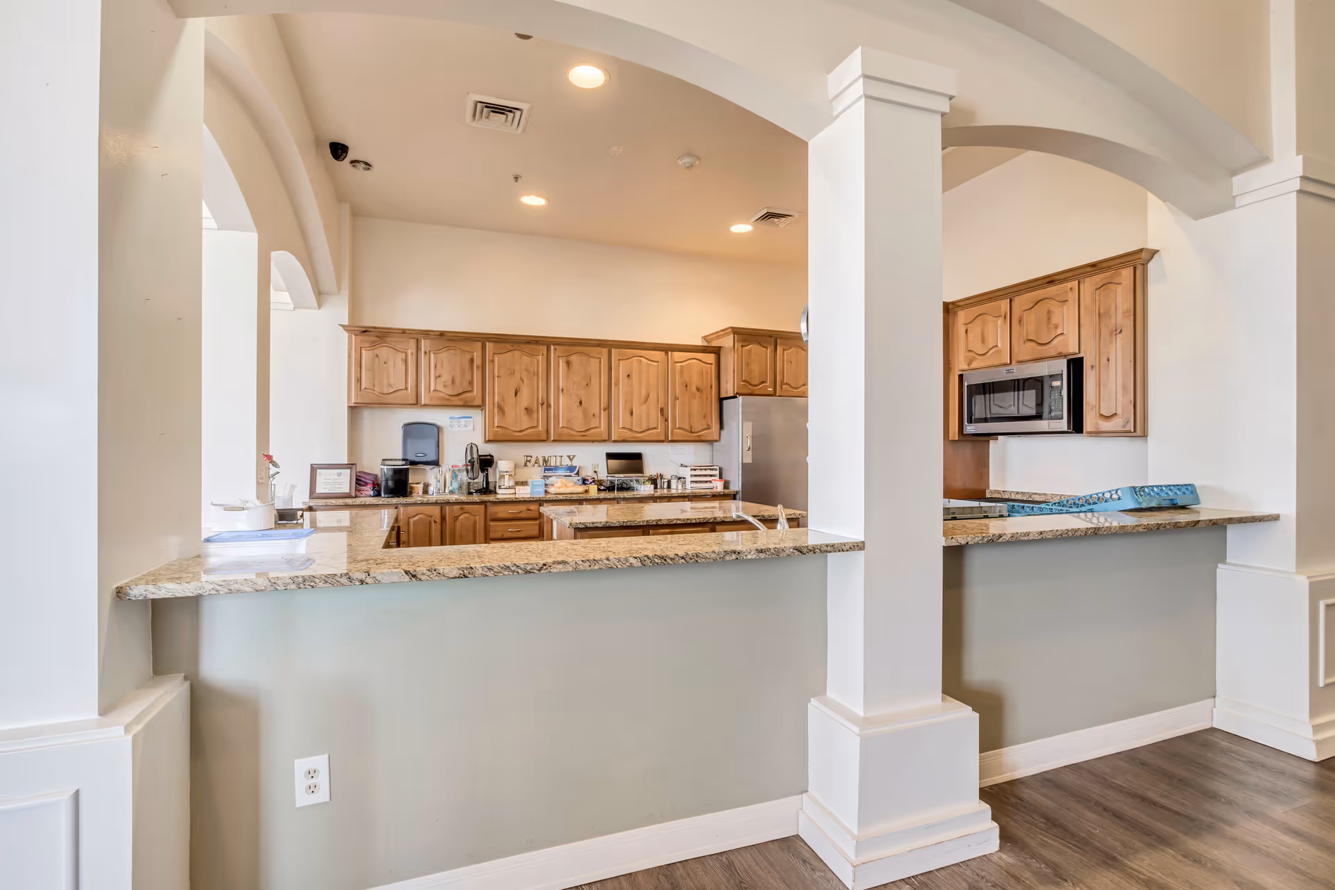 Interior view of a kitchen area with wooden cabinets, granite countertops, and built-in appliances including a microwave and refrigerator. The kitchen is separated from the adjacent room by a half wall with pillars and arches. The floor is wood, and the ceiling has recessed lighting.