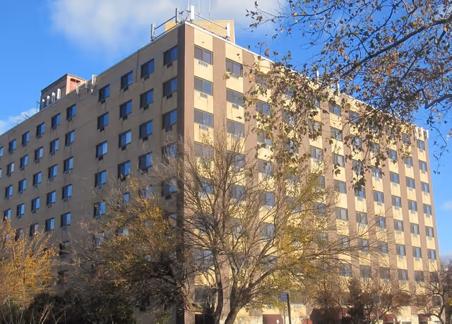 Tall brick nursing facility building with many windows, leafless trees, and blue sky.