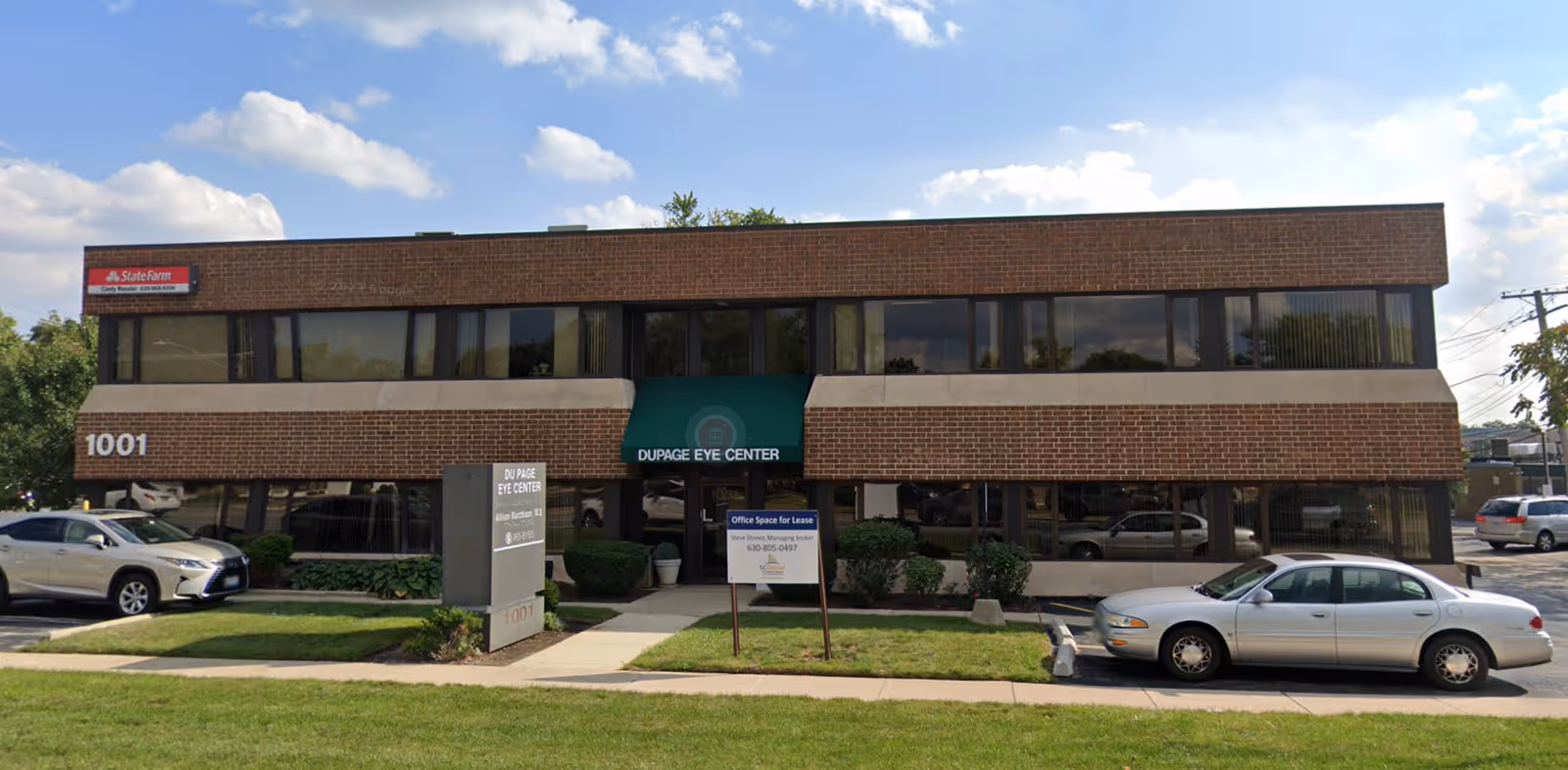 Two-story brick office building with large windows, green awning, and parked cars.