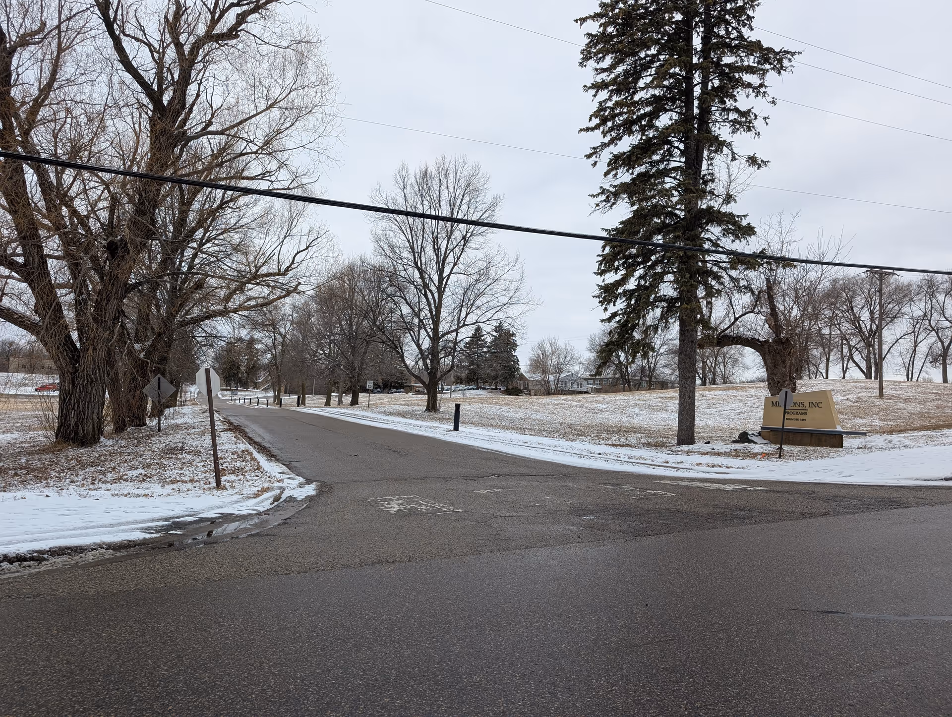Snow-dusted road and intersection lined with leafless trees and a grassy field with a facility sign on the right.