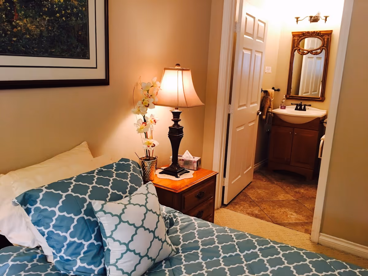 Cozy bedroom with patterned blue bedding, a bedside table and lamp, and an open door leading to a bathroom.