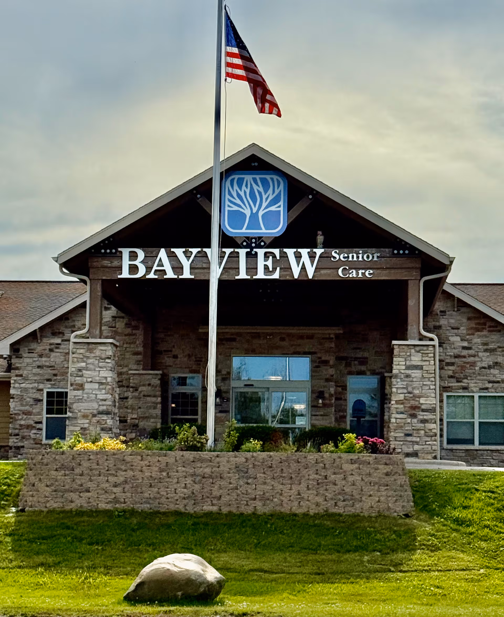 Front exterior view of Bayview Senior Care building with a stone facade, a covered entrance, an American flag on a flagpole in front, and a landscaped area with grass and a large rock.