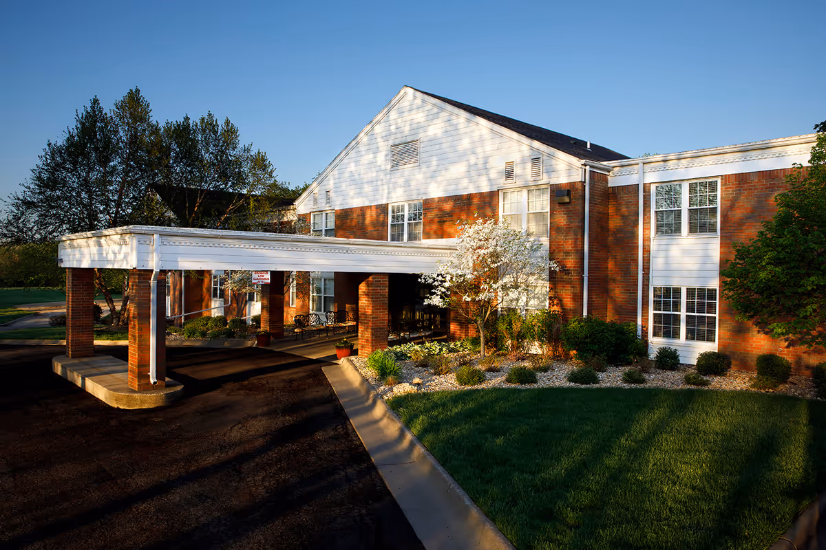 Exterior view of Lexington Park Assisted Living of Topeka building during daylight with a covered entrance supported by brick pillars, landscaped garden with bushes and a flowering tree, and a clear blue sky.