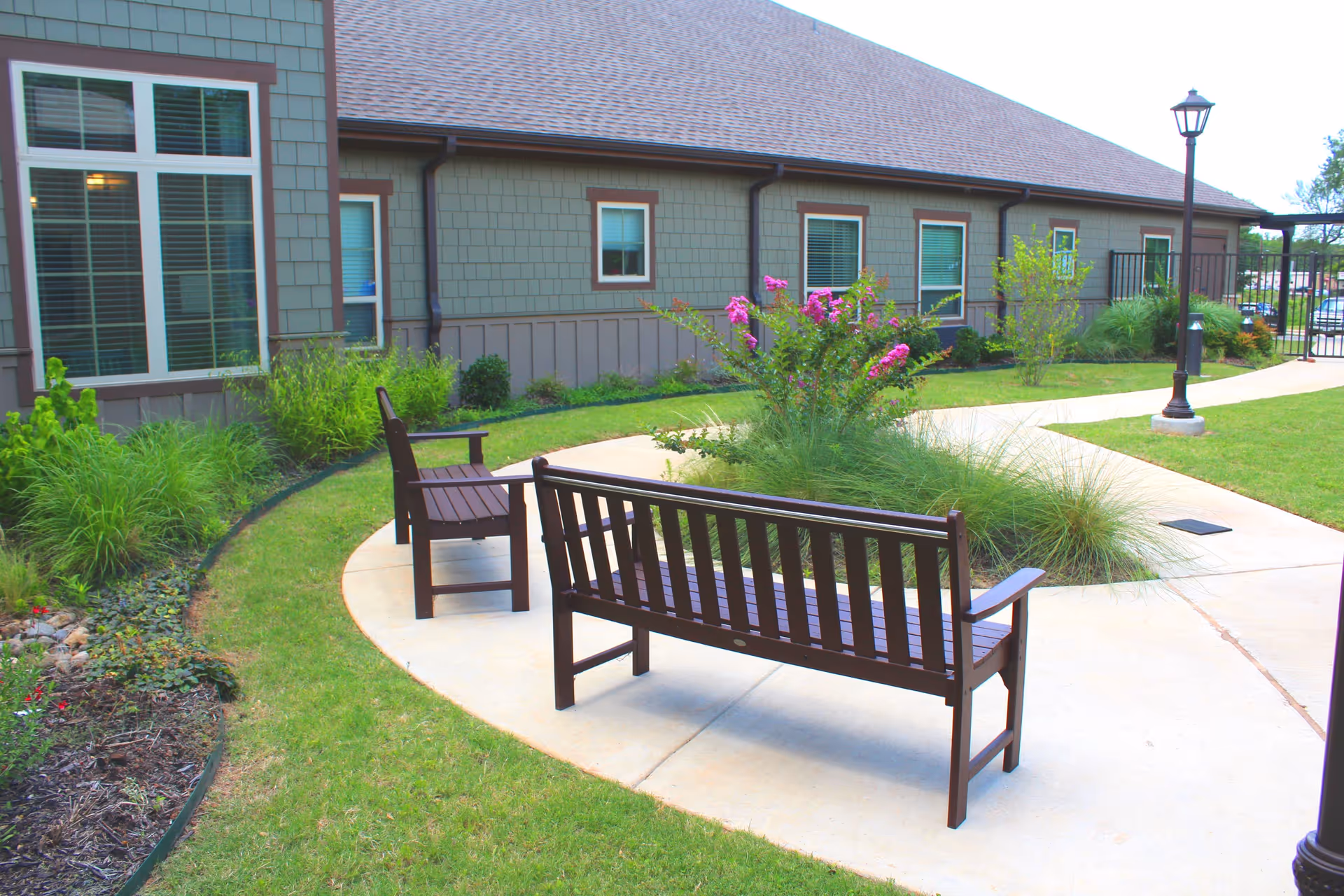 Outdoor garden area at Arbor Grace Guest Care Center featuring a curved concrete pathway with two wooden benches, surrounded by green grass, various plants, and flowers. The building exterior with windows and a lamp post is visible in the background.