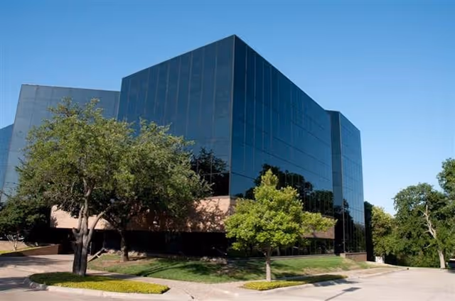 A modern glass-front building with surrounding trees and a driveway.