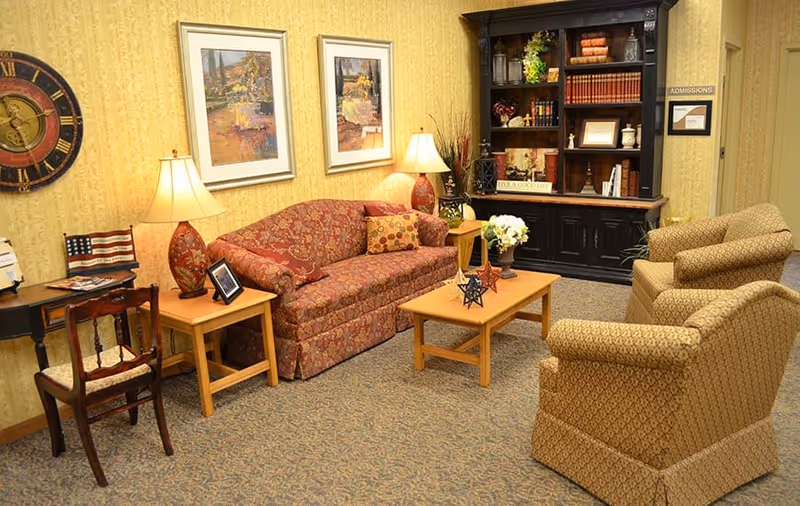 Cozy sitting room with a patterned sofa, two upholstered armchairs, coffee table, lamps, and a bookcase against a wallpapered wall.