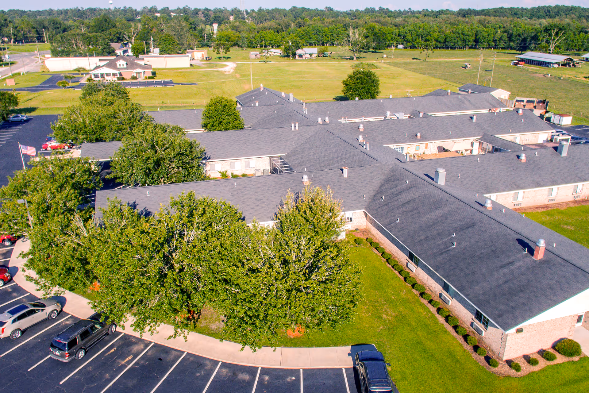 Aerial view of a large single-story building with a dark roof surrounded by green trees and grass. There is a parking lot with several cars in front of the building. The area around the building is open with fields and more trees in the background.