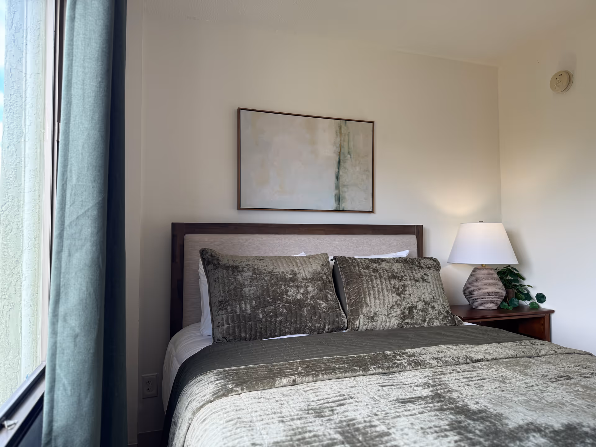 A neatly made bed with a textured greenish-gray bedspread and matching pillows in a bedroom. There is a wooden nightstand with a lamp and a small plant on the right side of the bed. A large abstract painting hangs on the wall above the bed. A window with green curtains is visible on the left side.