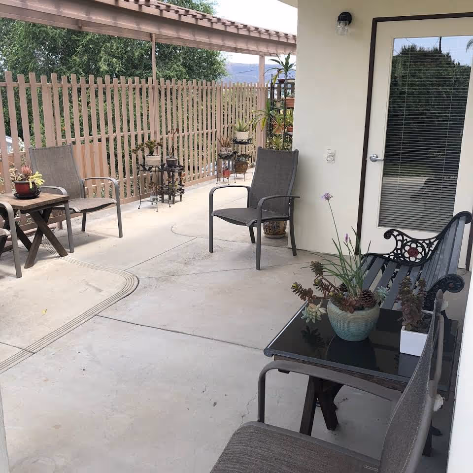 Covered patio with outdoor chairs, side tables, a bench and several potted plants next to a glass door.
