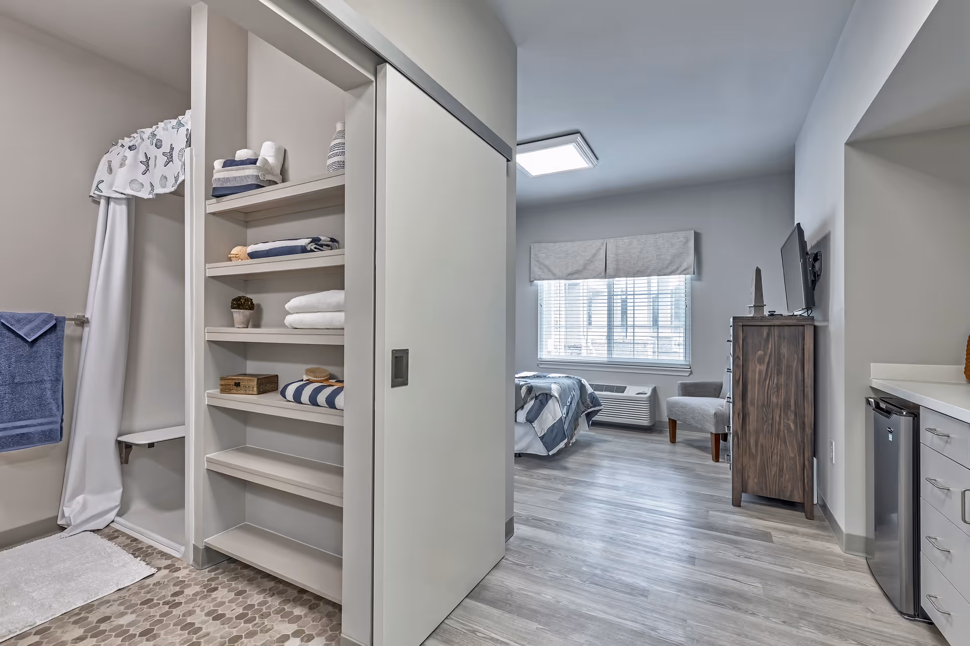 Interior view of a senior living apartment: open closet, bed, window, and kitchenette.