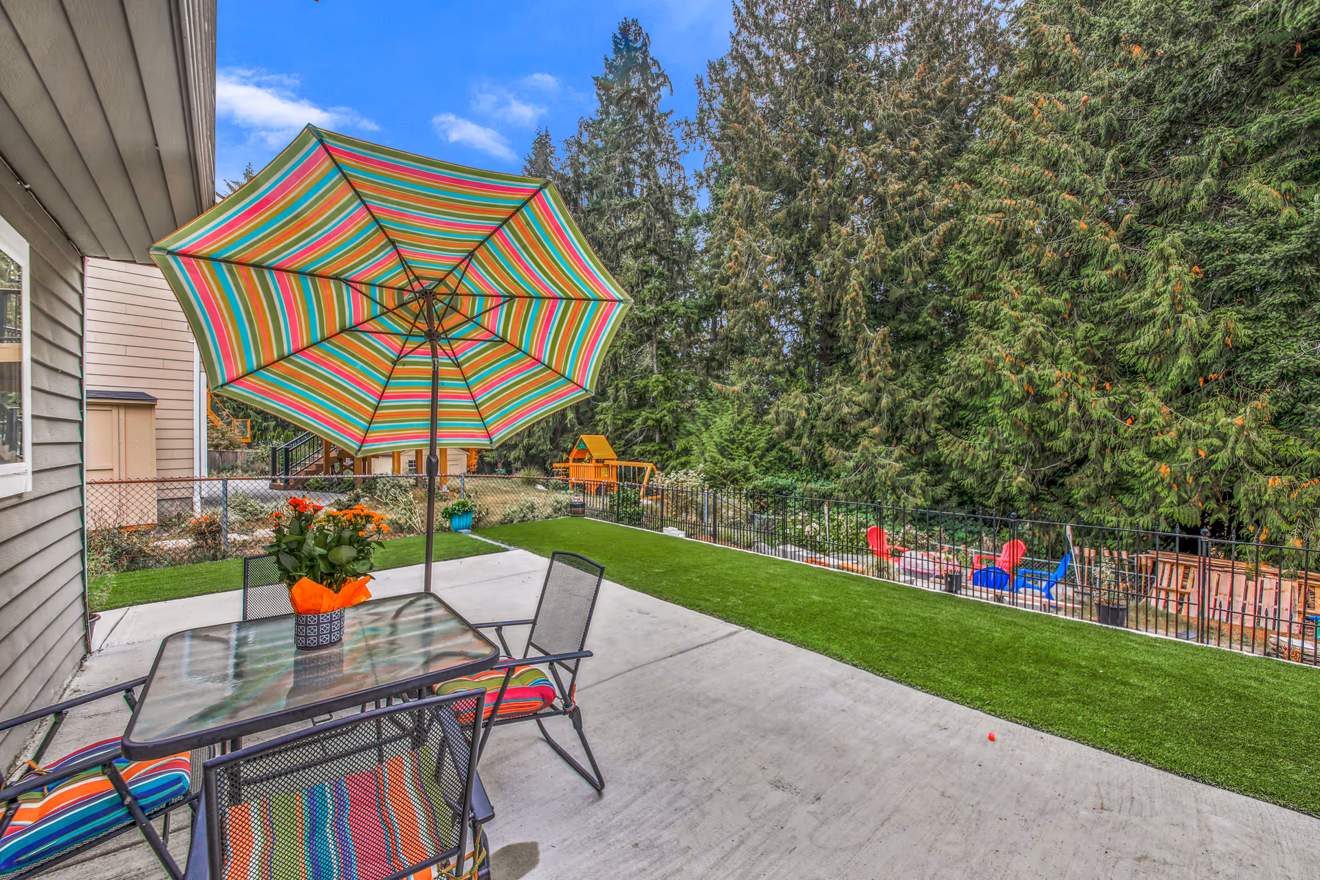 Outdoor patio with striped umbrella, glass table, chairs, and a fenced yard surrounded by trees.