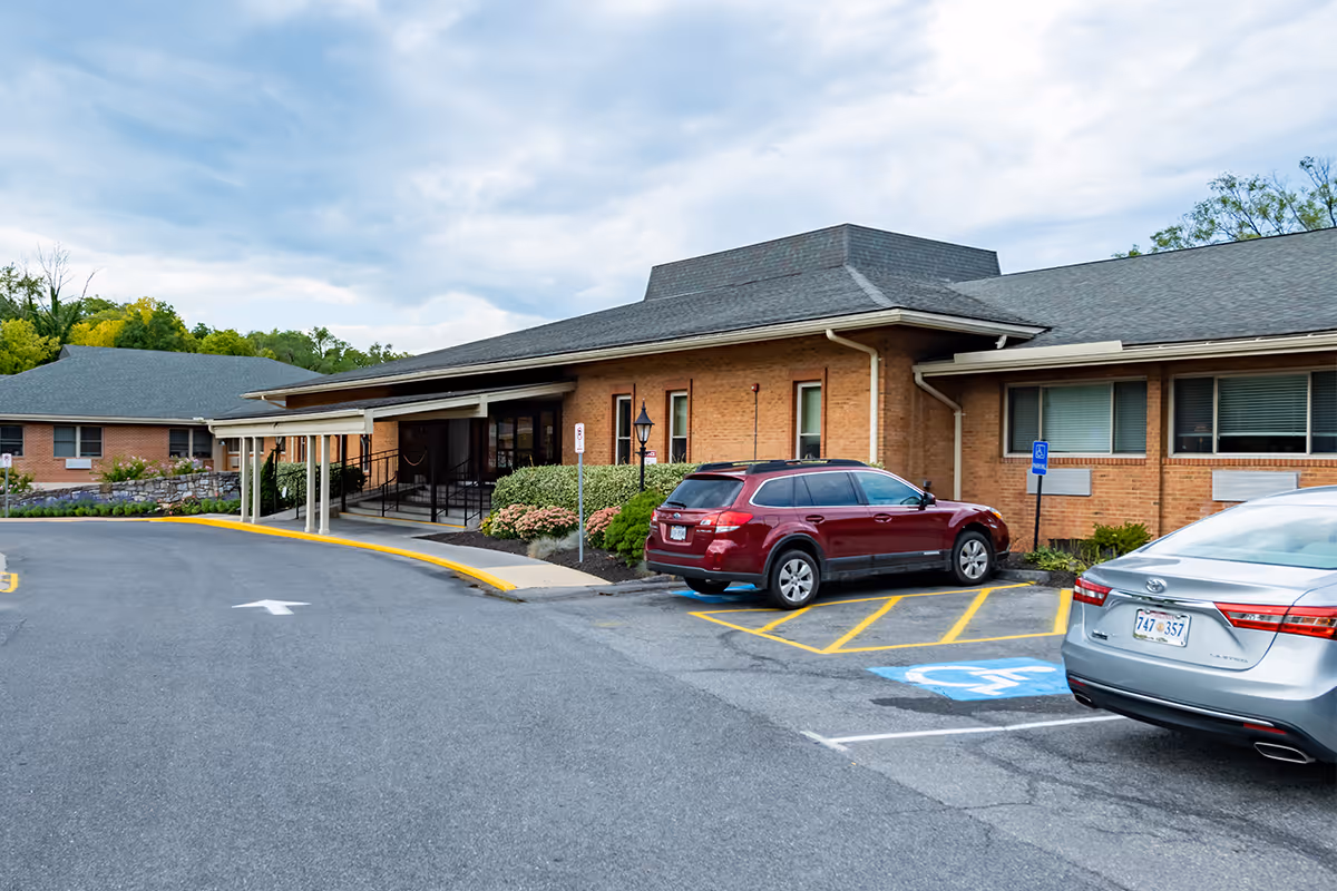 Brick senior living facility exterior with a driveway, entrance canopy, and parked cars.