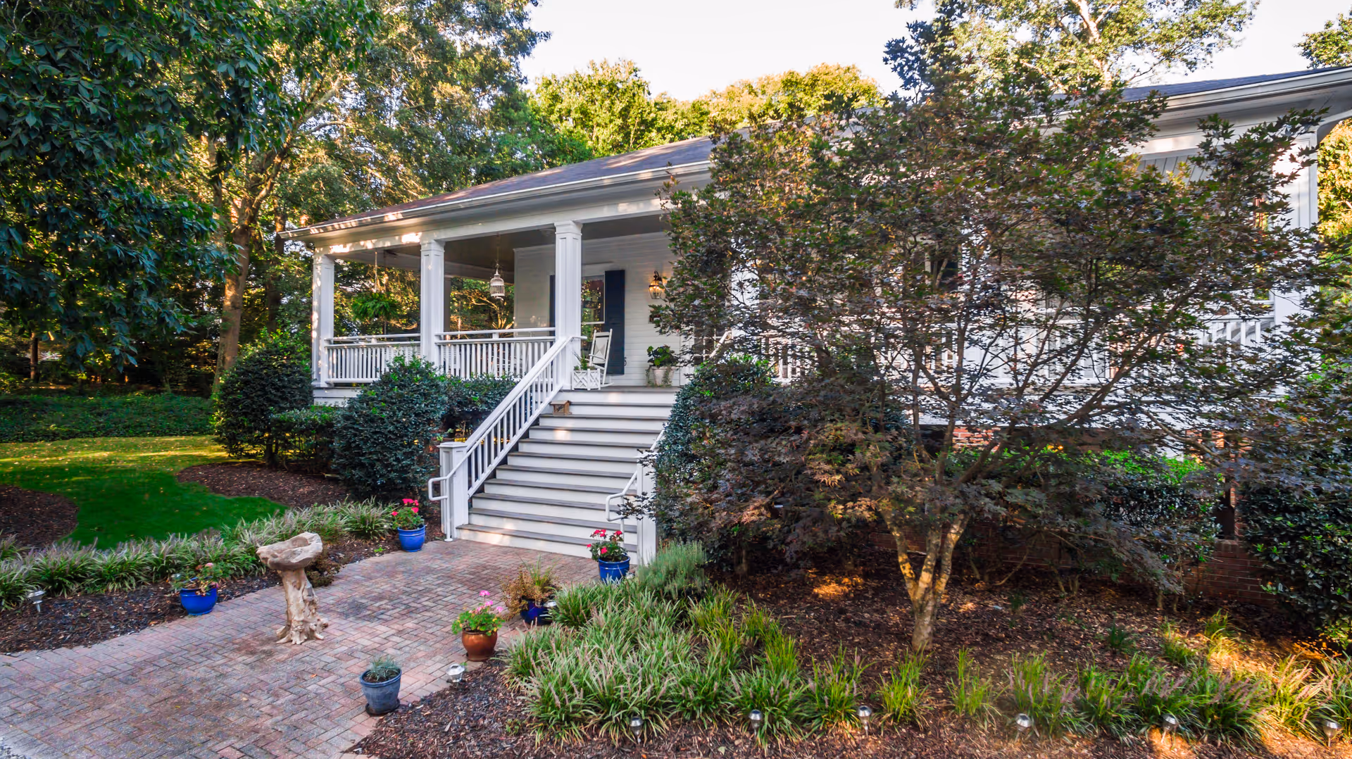 A white front porch with steps and railing overlooks landscaped garden and trees.