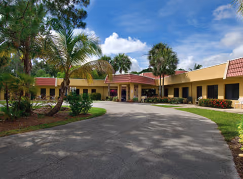 Driveway leading to the entrance of a single-story senior living building with palm trees and landscaping under a blue sky.
