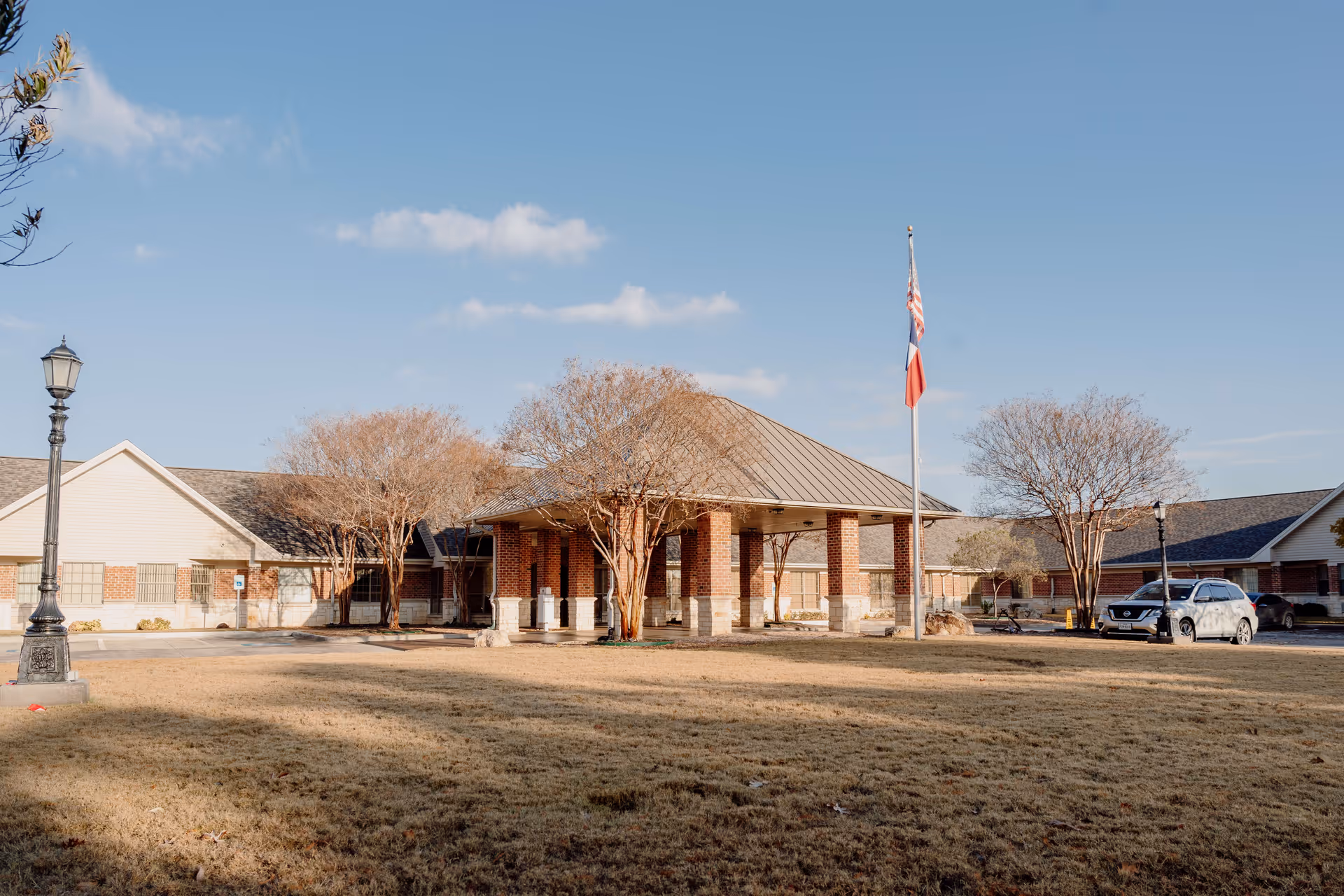 Exterior view of Azle Manor Nursing Home building with a covered entrance supported by brick columns, a flagpole with the American and Texas flags, leafless trees, a street lamp, and a parked car on a clear day.