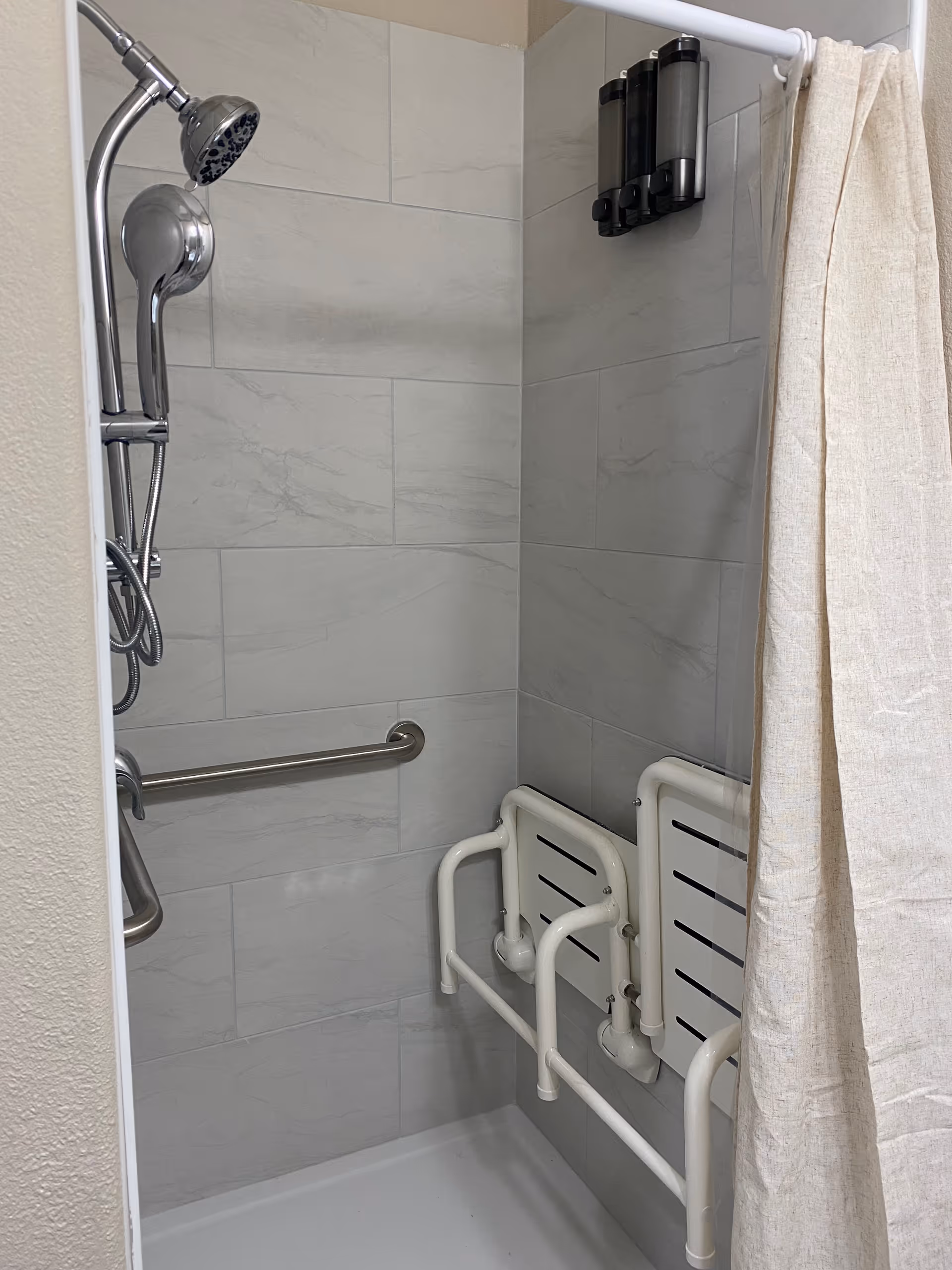 Accessible shower with handheld showerhead, grab bars, and fold-down seat in a tiled bathroom.
