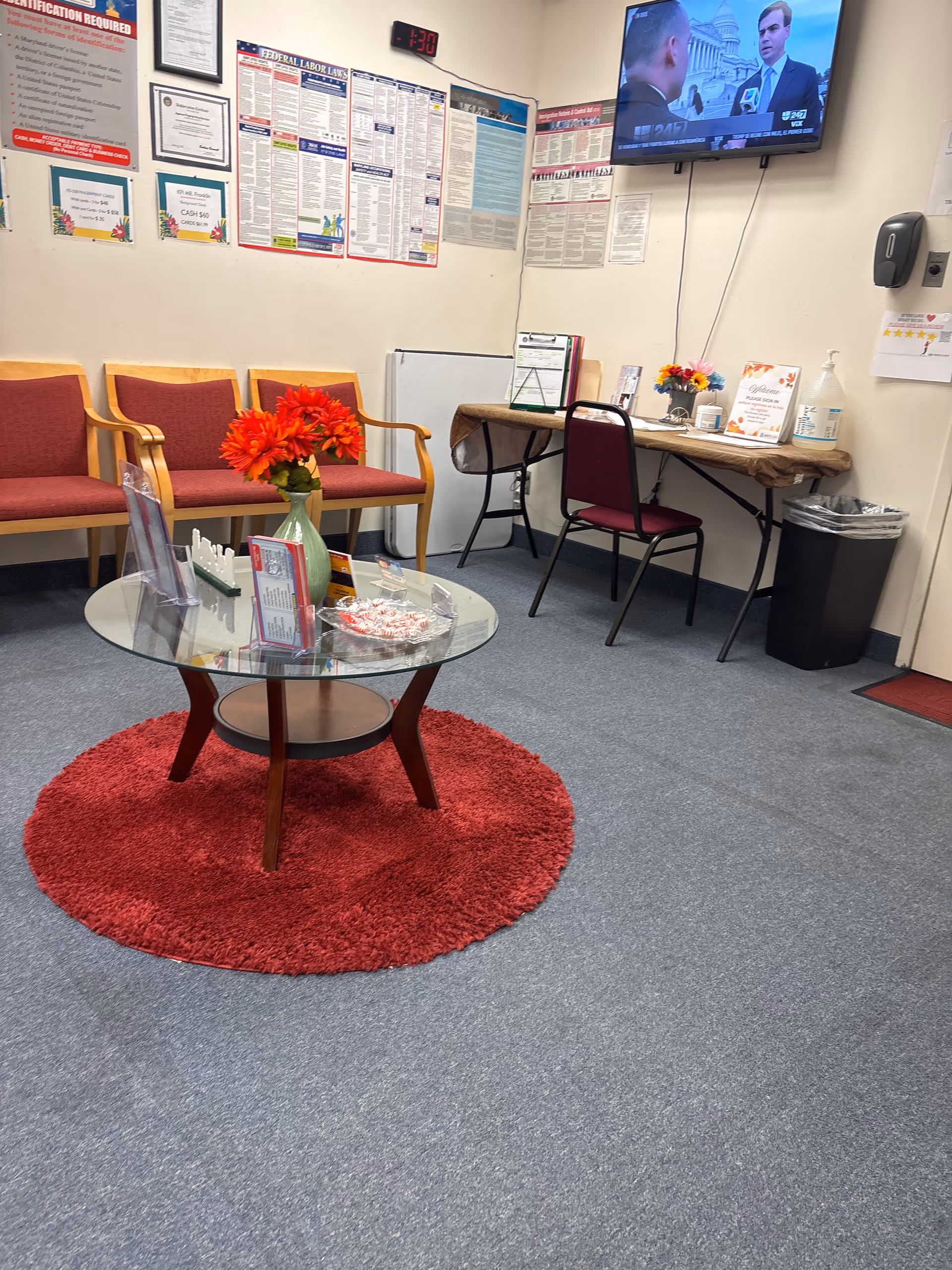 Indoor waiting area with red chairs, glass coffee table, wall posters, and a mounted TV.