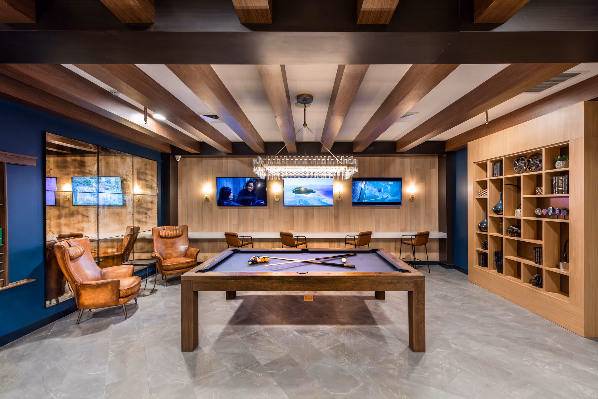 A stylish game room featuring a pool table with cues and balls arranged on it. The room has wooden ceiling beams, a modern chandelier above the pool table, and three wall-mounted TVs displaying different content. There are two brown leather armchairs on the left side near a large mirror, and a wooden shelving unit with decorative items on the right. Four chairs are positioned along a counter beneath the TVs.