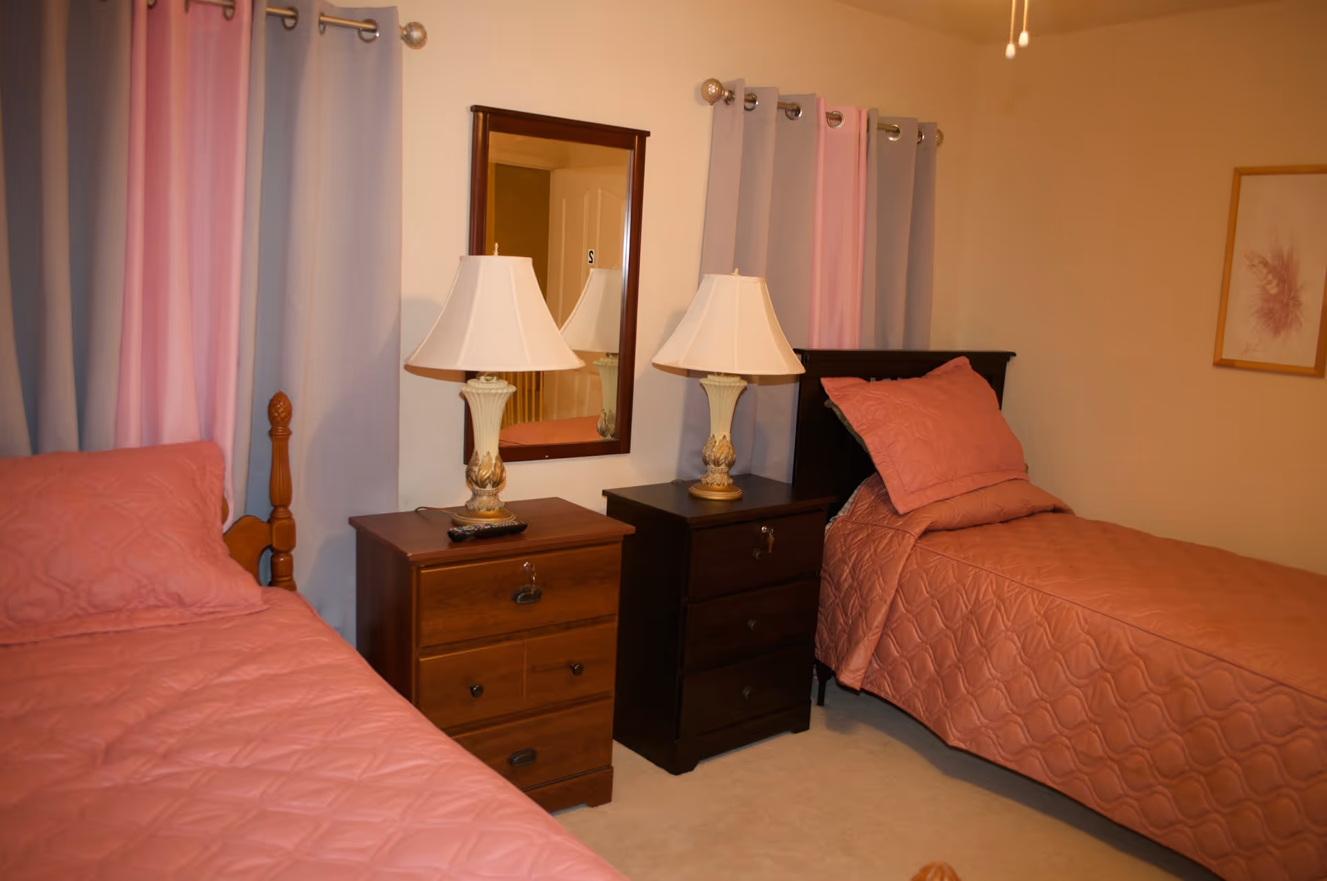 A bedroom with two twin beds covered in pink quilts, two bedside tables with lamps, a mirror, and curtains.