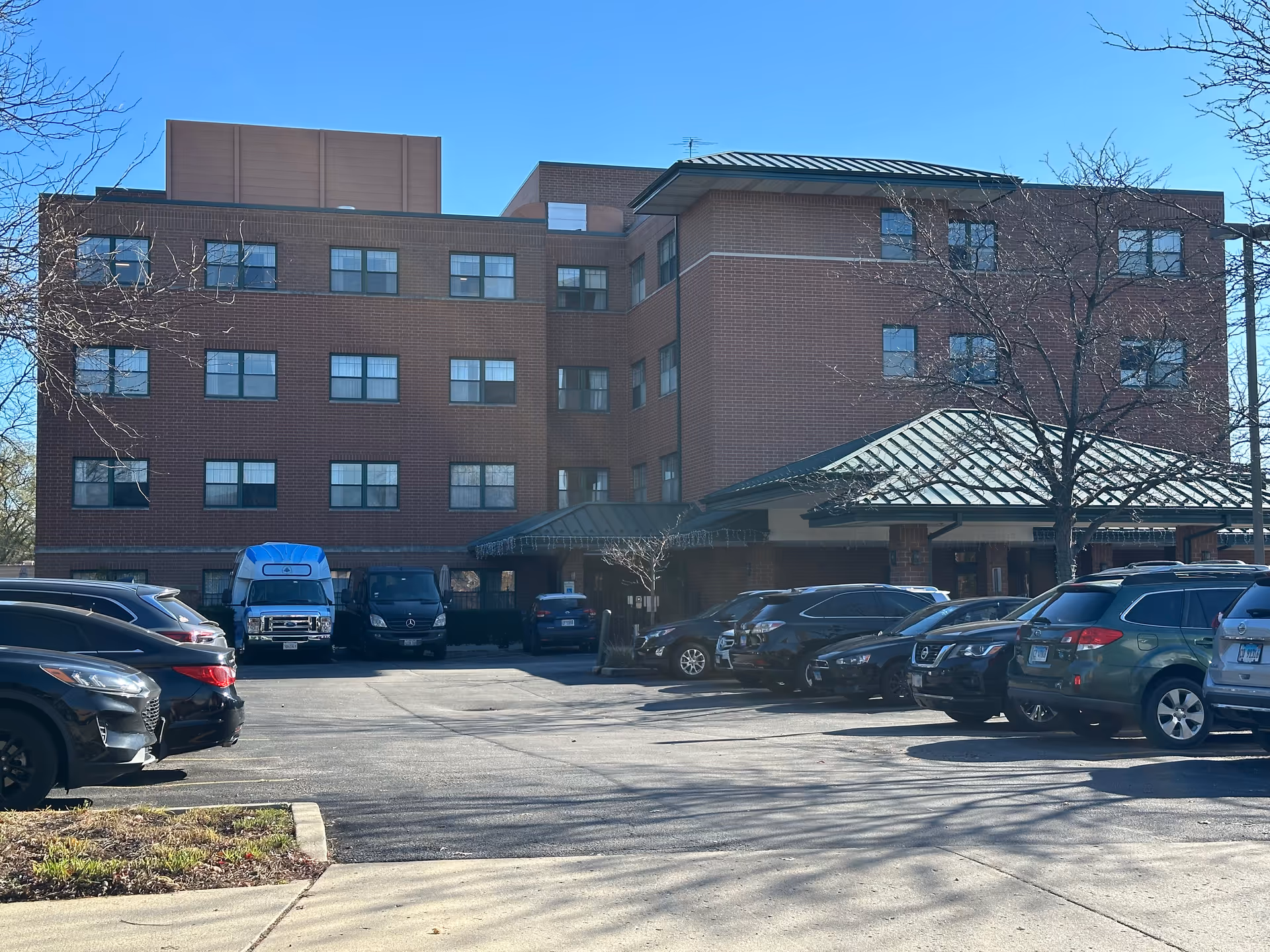 Exterior view of a four-story brick building with multiple windows and a green metal roof over the entrance. Several cars are parked in the parking lot in front of the building under a clear blue sky. Leafless trees are visible near the building.