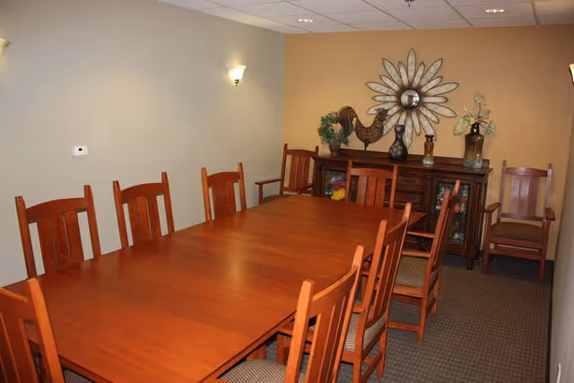 A dining room with a long wooden table surrounded by wooden chairs with cushioned seats. The walls are painted beige and light yellow, with two wall-mounted light fixtures. At the far end of the room, there is a wooden sideboard decorated with a large flower-shaped mirror, a rooster figurine, and vases with plants.