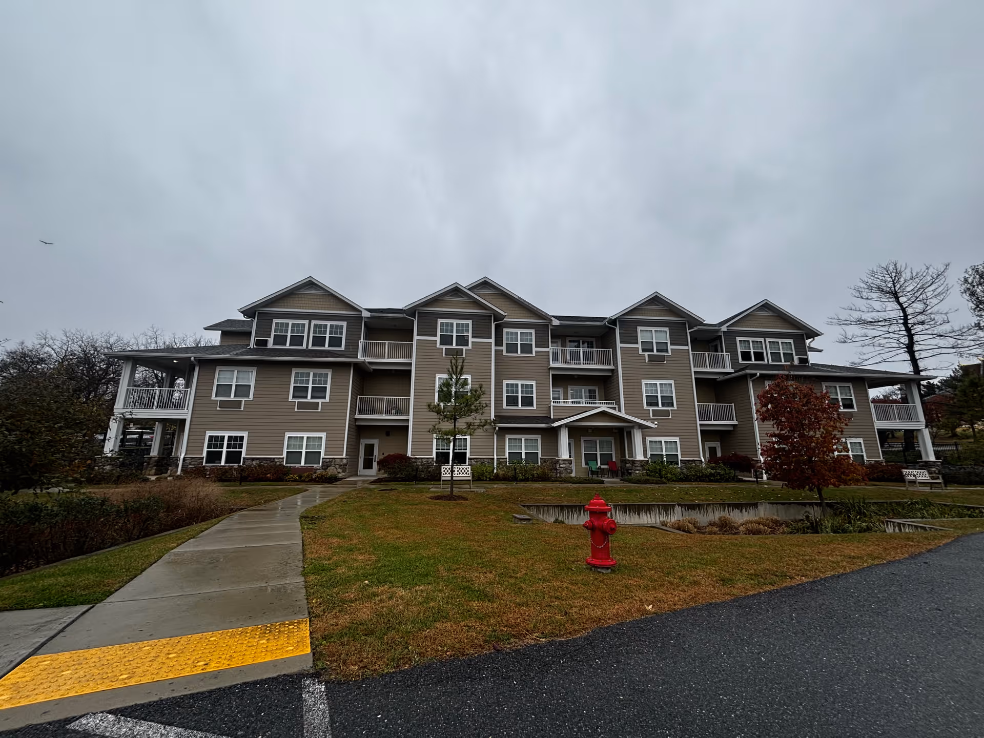 Front view of a three-story beige retirement apartment building with balconies, lawn, a small pond and a red fire hydrant under a cloudy sky.