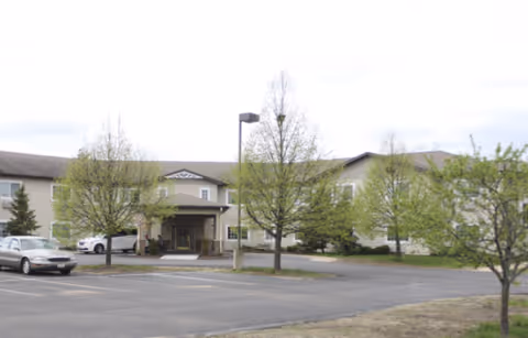 Front exterior view of a two-story senior living facility named Oak Gardens Place with a covered entrance, several small trees with green leaves, and a parking lot with a few cars.