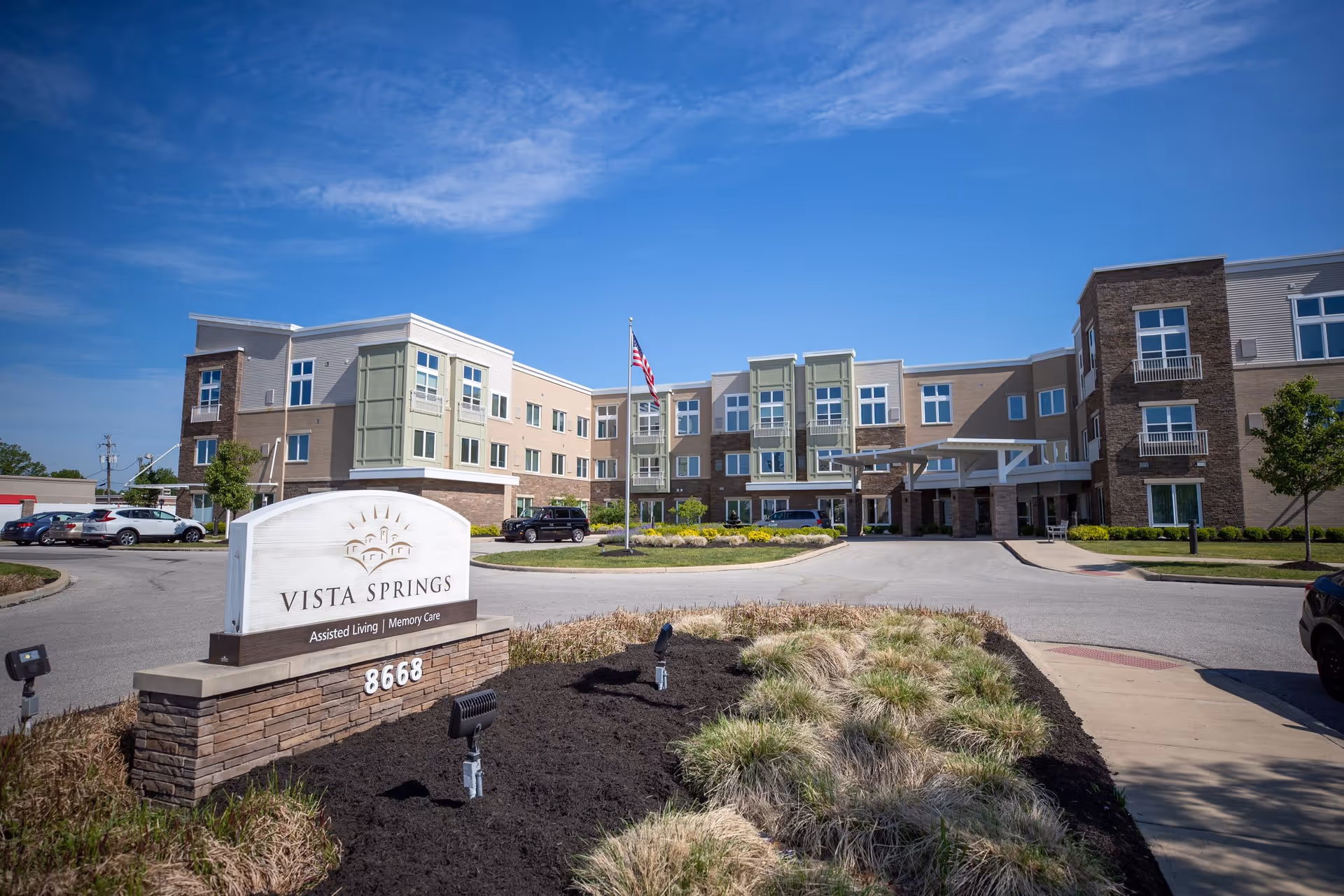 Exterior view of Vista Springs Greenbriar Village, a multi-story assisted living and memory care facility with a landscaped entrance, a sign displaying the facility name and address, and an American flag in front under a clear blue sky.