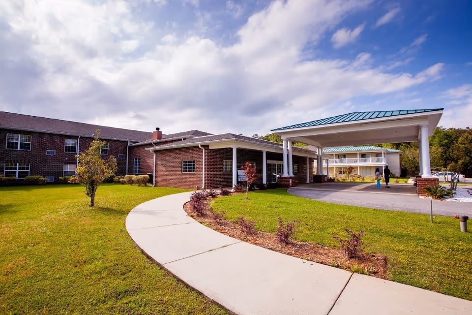 Front exterior of a brick senior living facility with a covered entrance, curved walkway, and landscaped lawn under a partly cloudy sky.